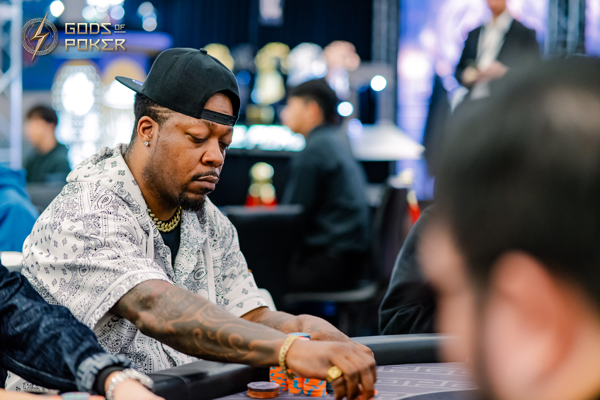 Man with backward cap and gold jewelry intently playing poker at a casino table.