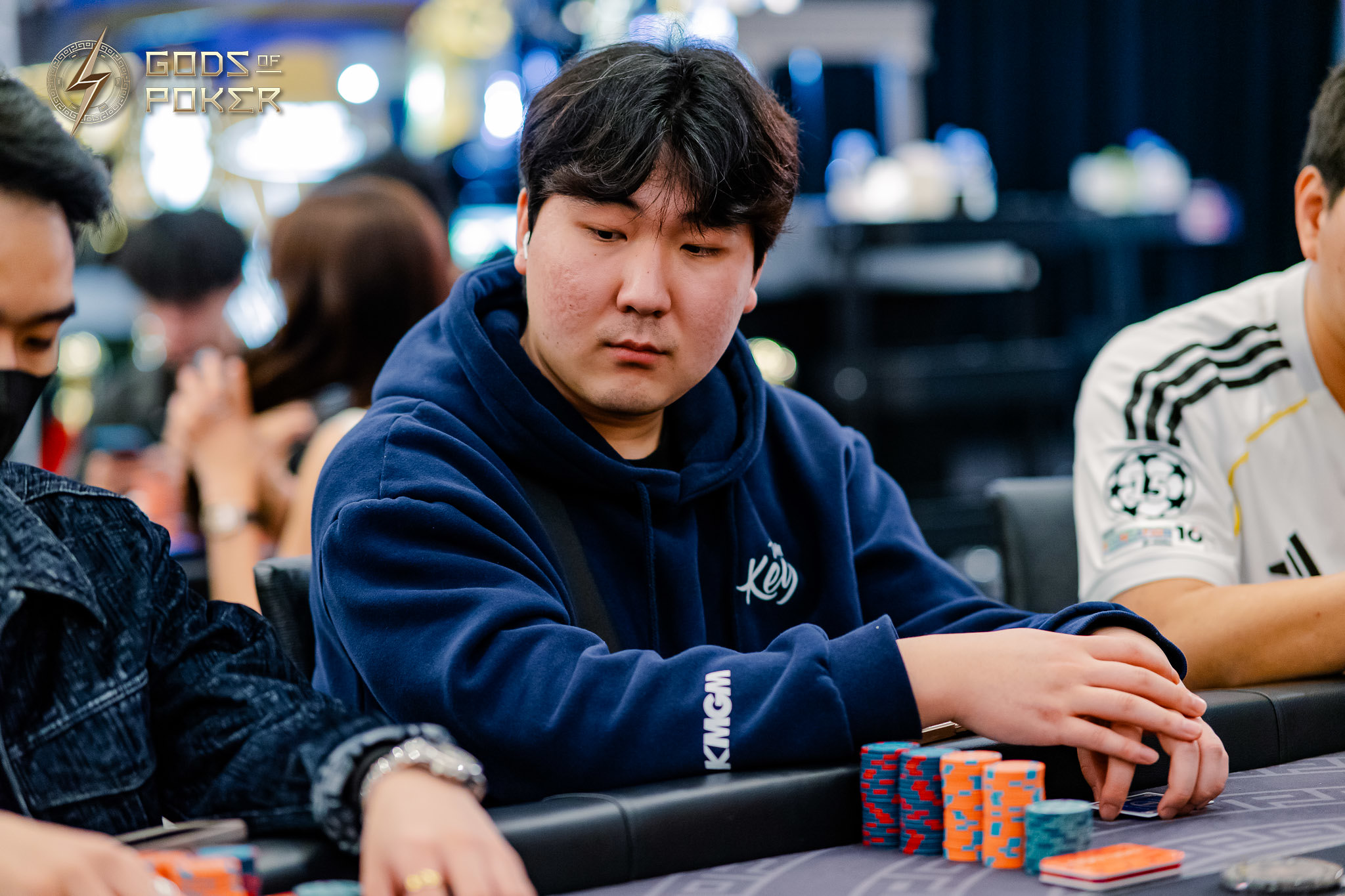 An Asian man in a blue hoodie, looking contemplative at a poker table with chips.