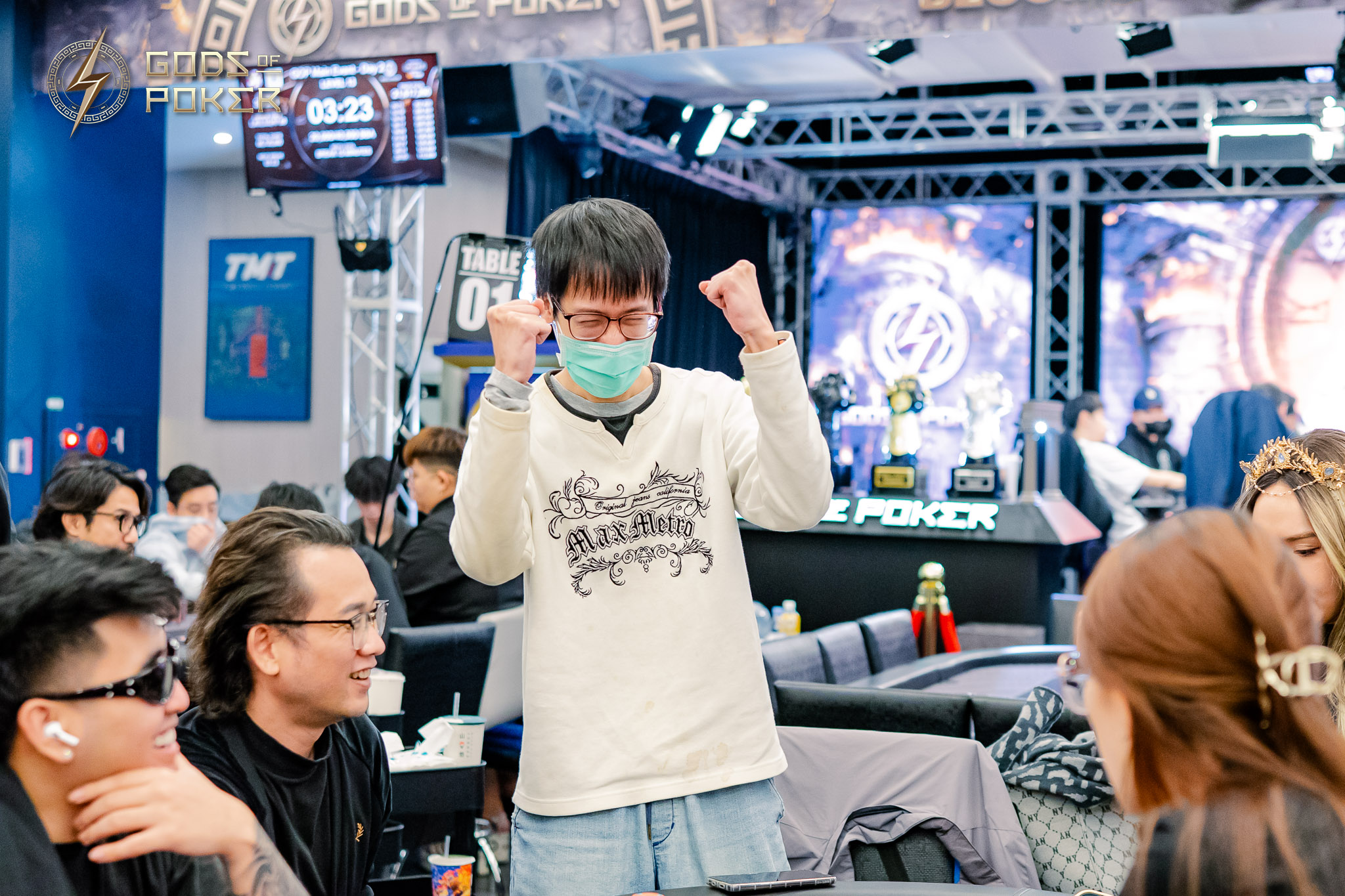 Man in surgical mask celebrates with clenched fists at a busy poker tournament.