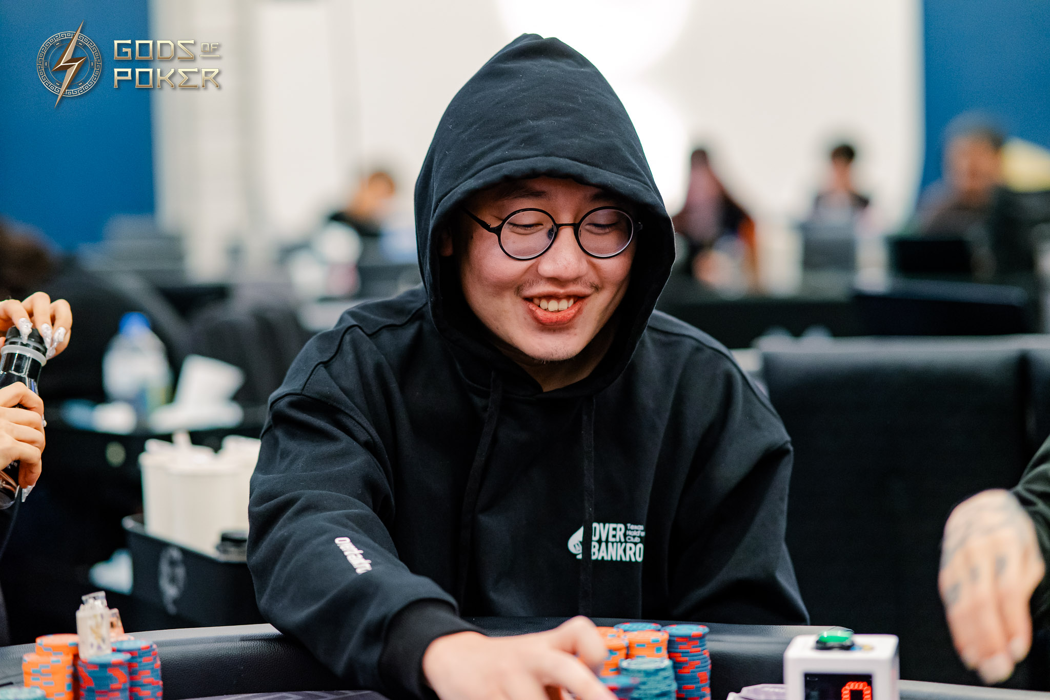 Young man in a black hoodie and glasses smiles while playing poker.