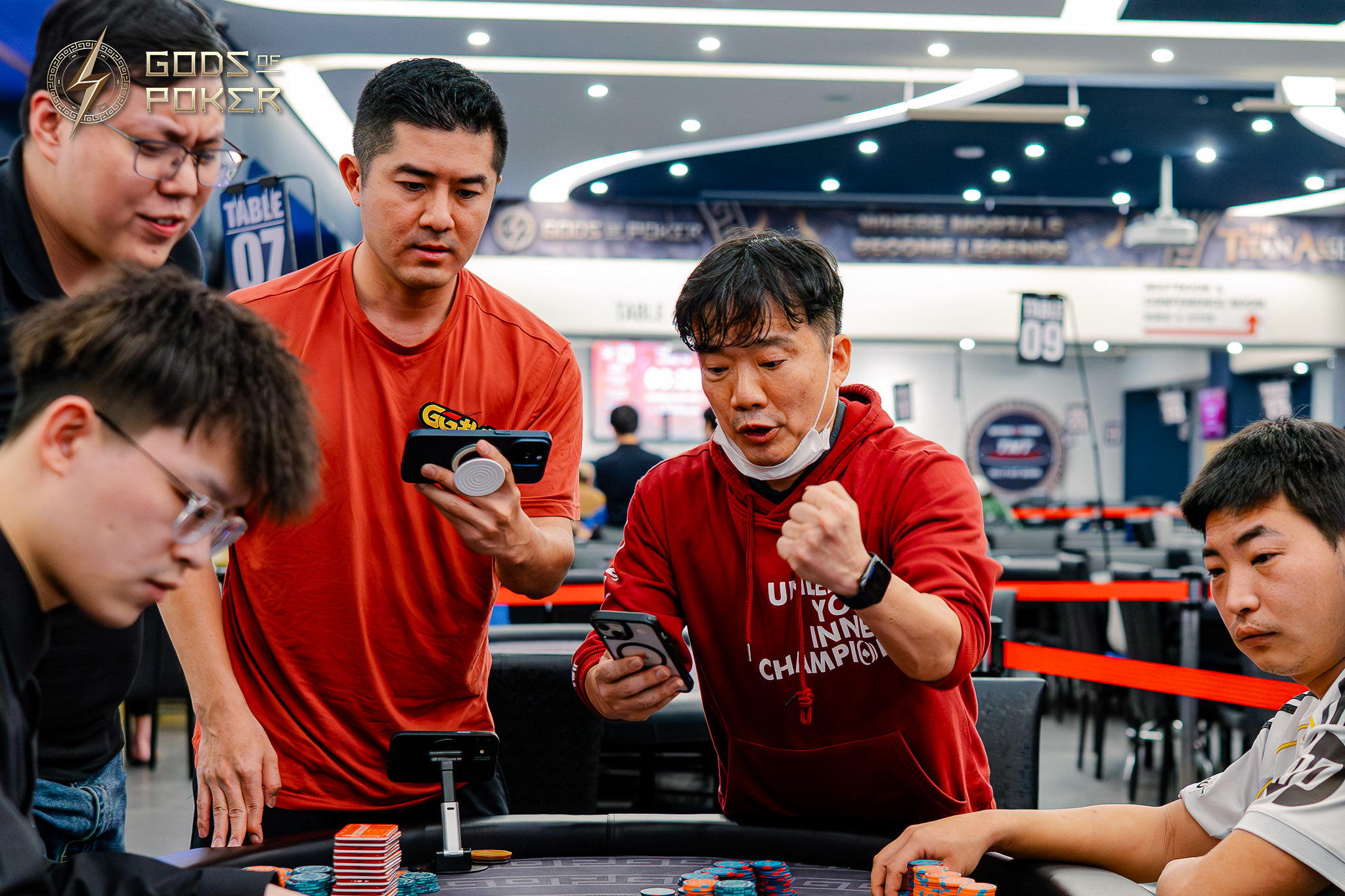 Men observe a poker game, one animatedly reacts while holding a phone.