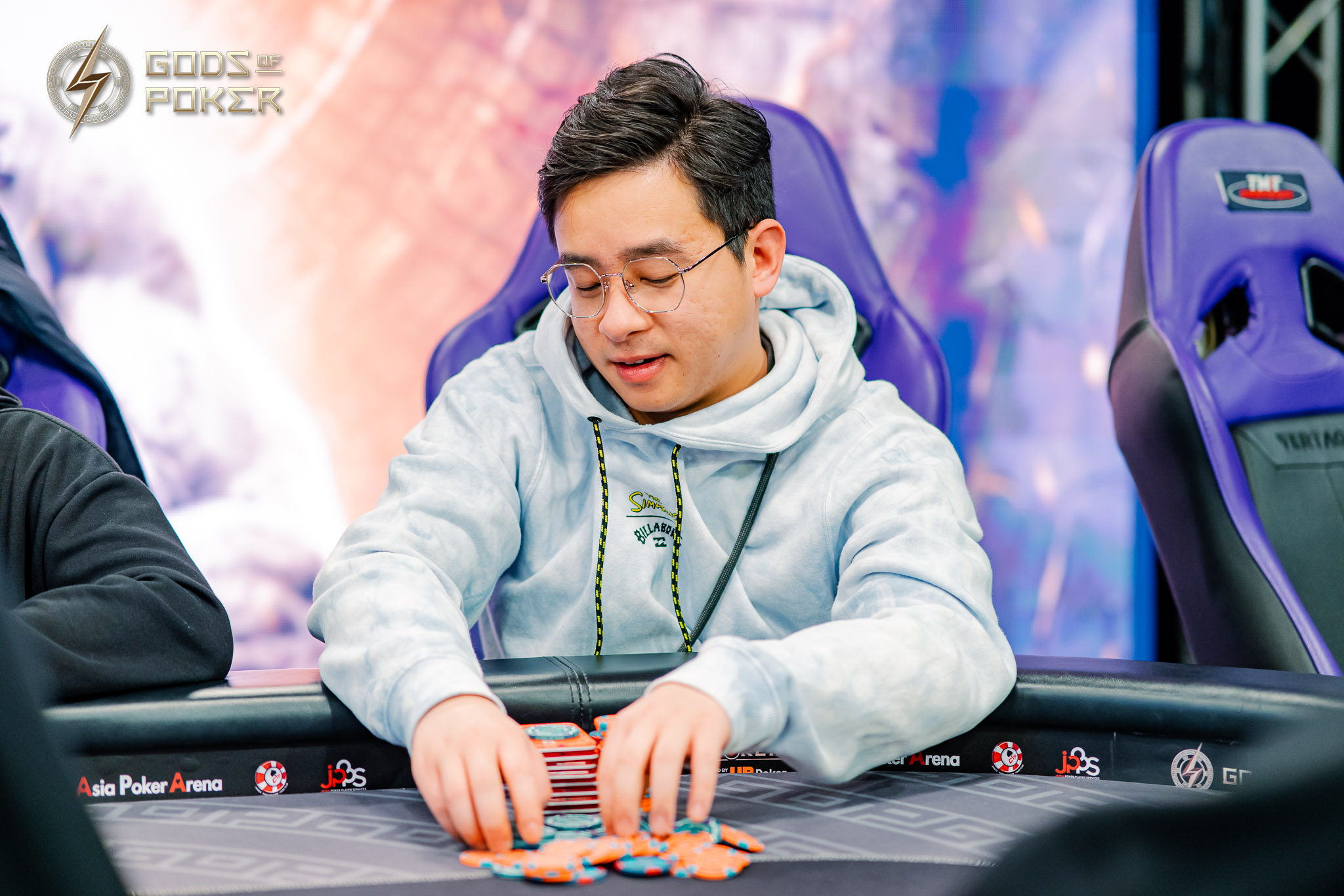 A man in glasses and a light blue hoodie moves poker chips at a table.