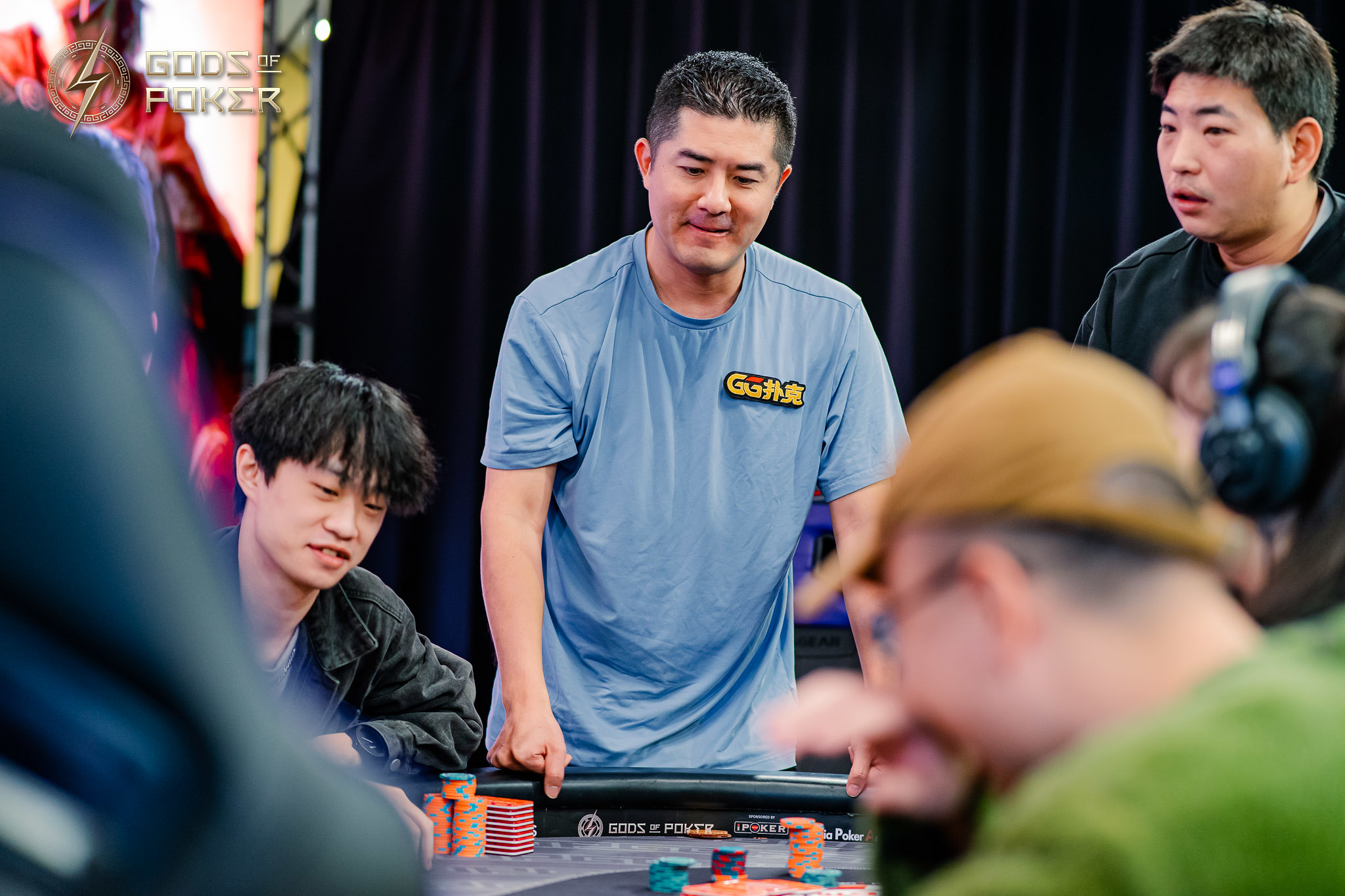 A man in a blue shirt standing at a poker table, surrounded by other players.