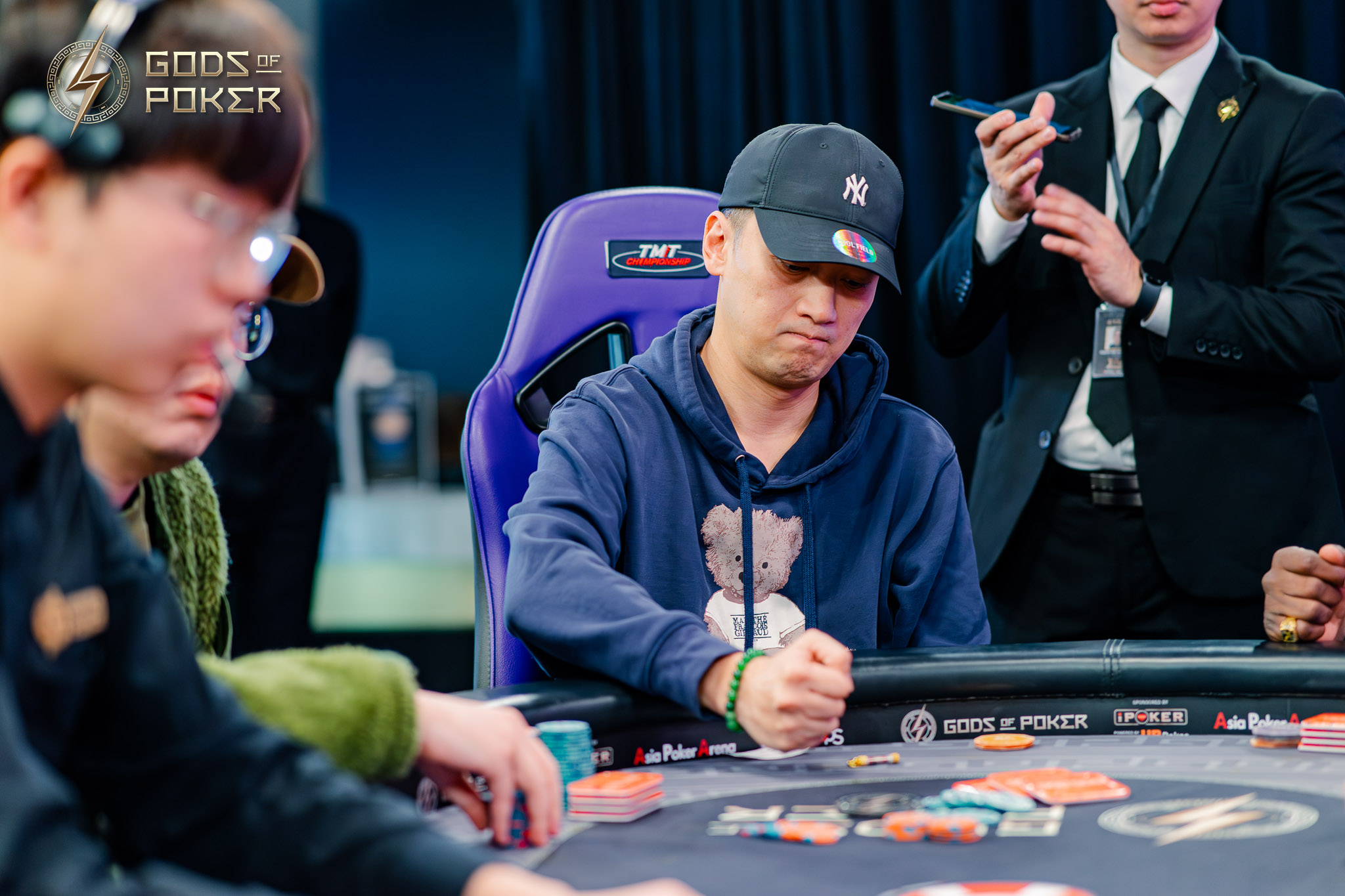 Man in black cap and blue hoodie deeply considers his hand at a poker table.