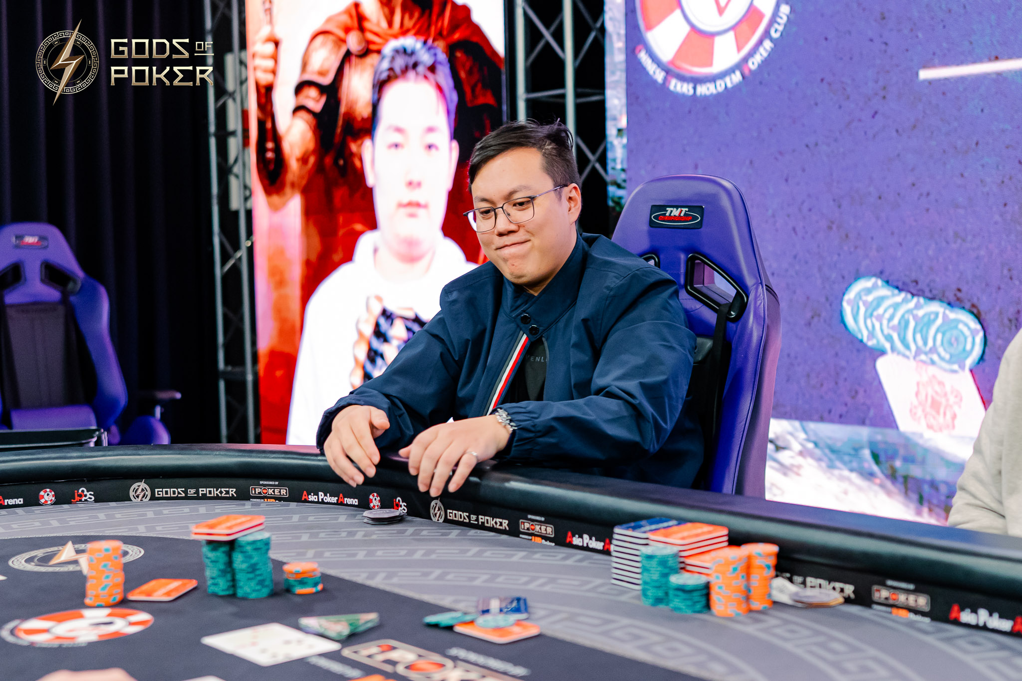 A man in a blue jacket sits at a poker table with chips and cards.