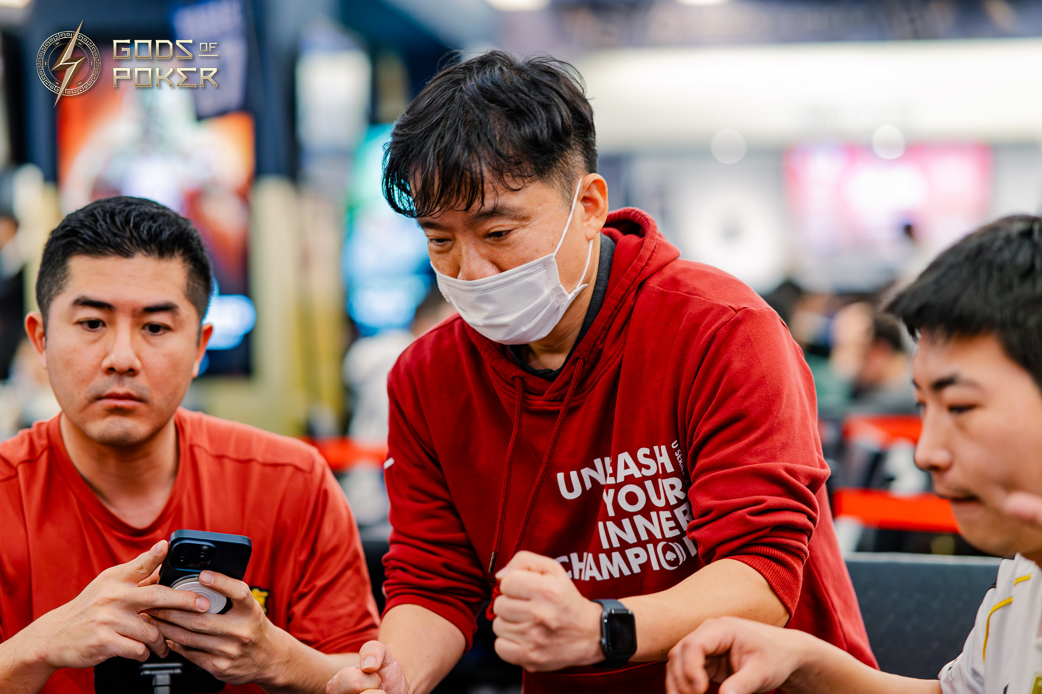 Man in red hoodie and mask intensely focuses during a poker game at a table.