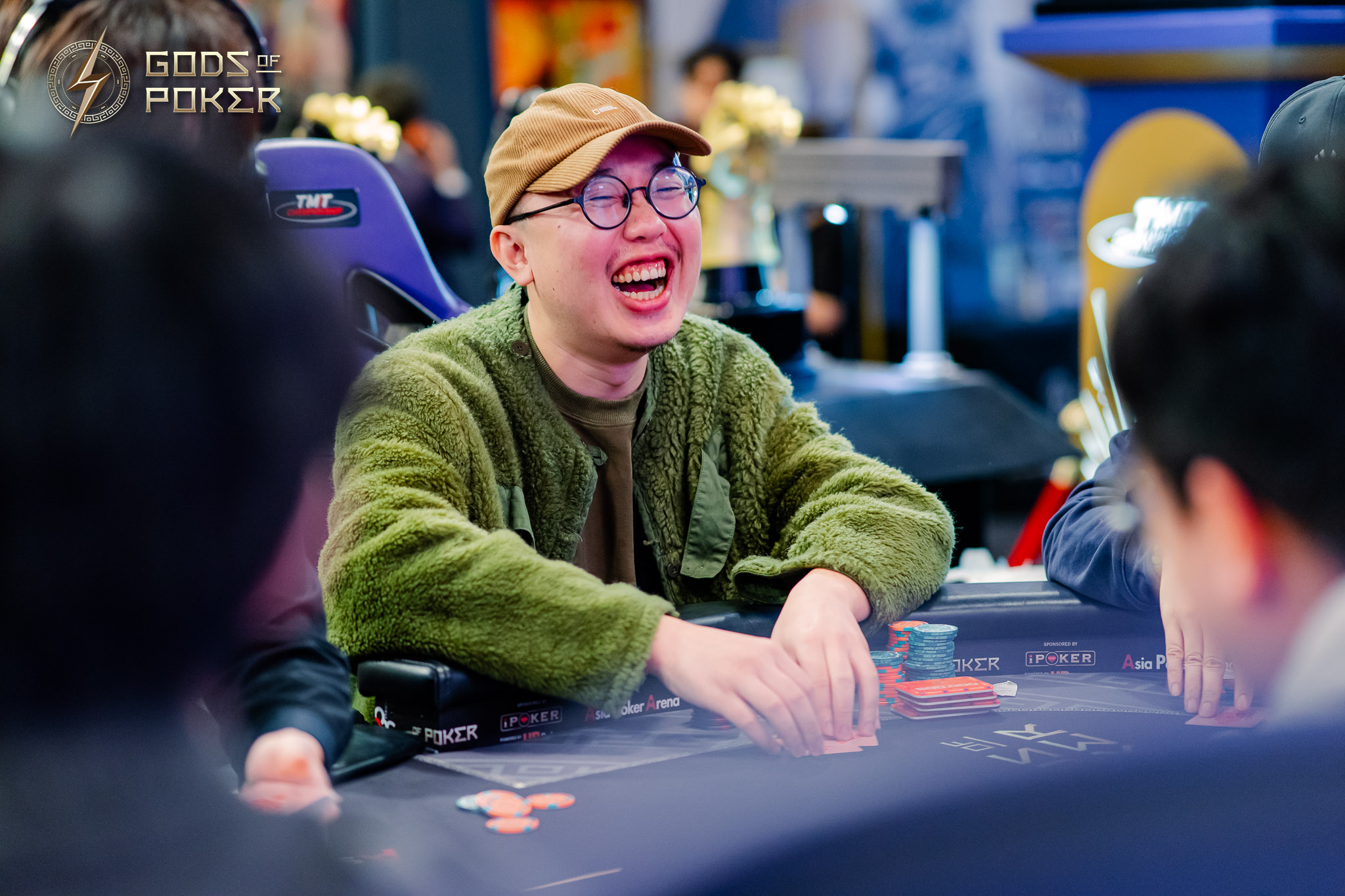 A man in a green jacket and cap laughs joyfully at a poker table with chips.