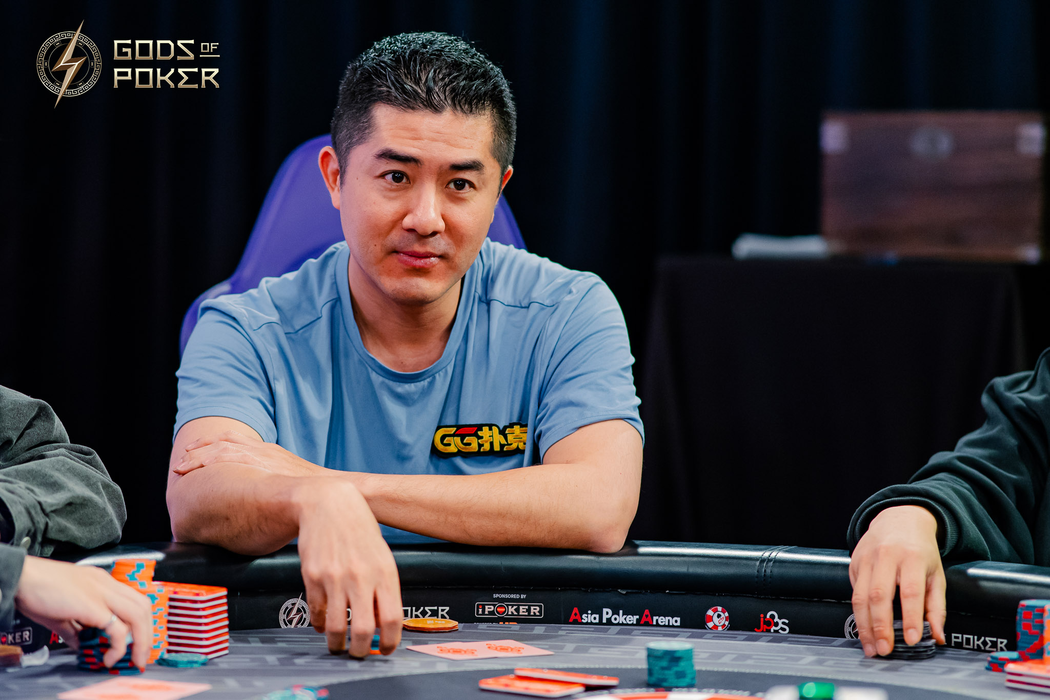 Asian man with a serious expression sits at a poker table, wearing a blue 'Game' shirt.
