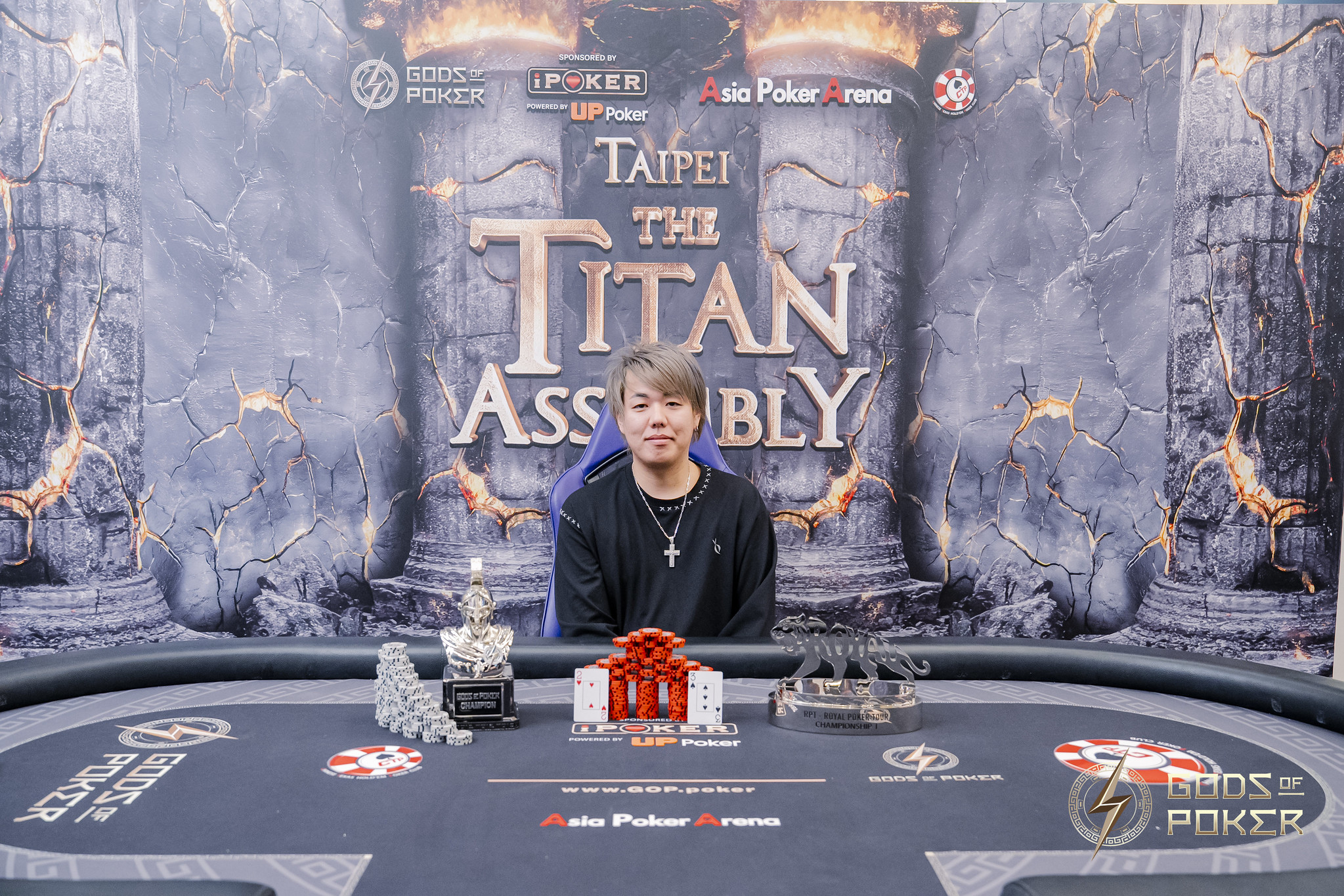 Young man smiles behind poker table adorned with trophies at 'The Titan Assembly'.