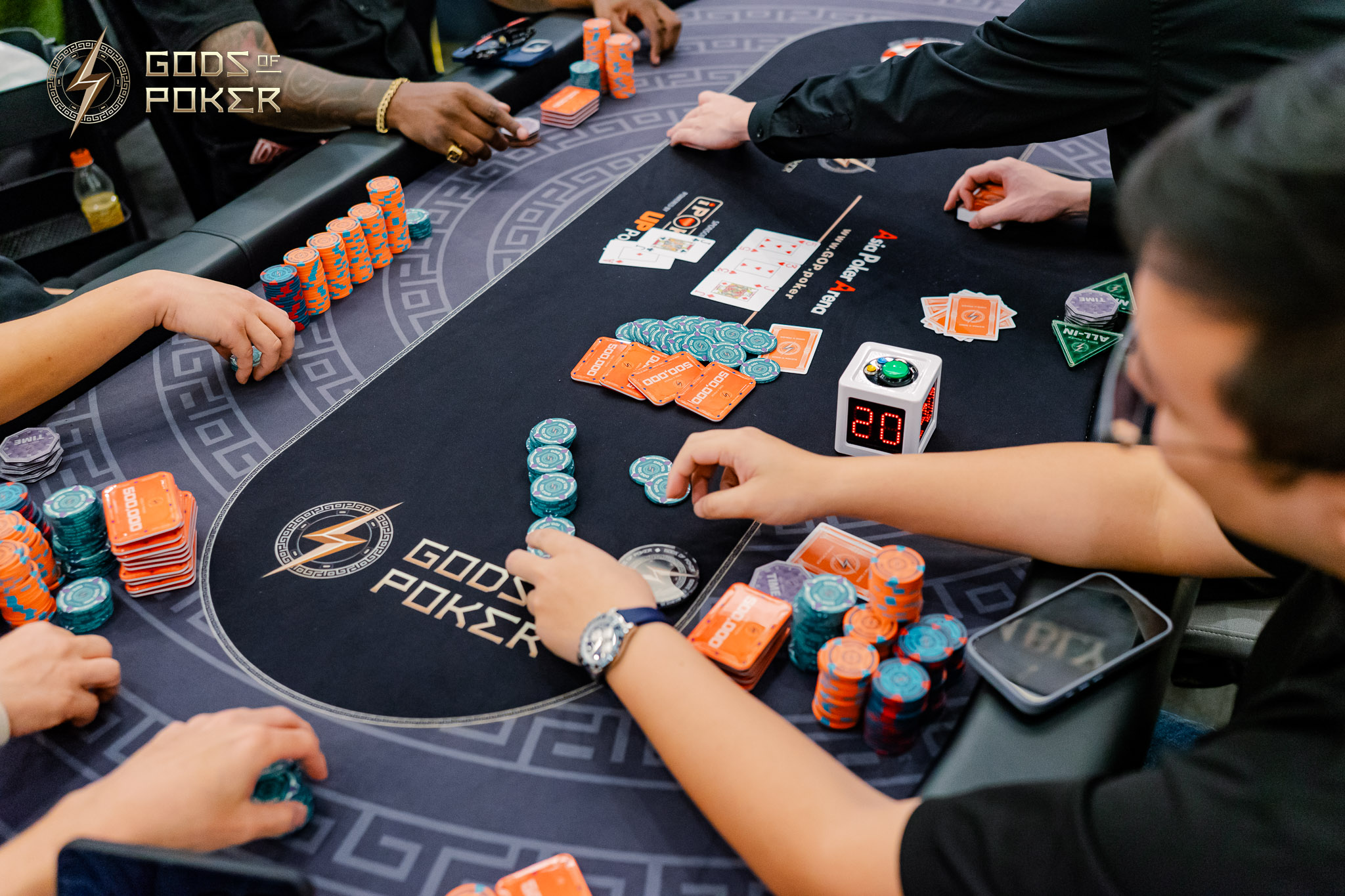 Players' hands arrange poker chips and cards on a black 'GODS POKER' table.