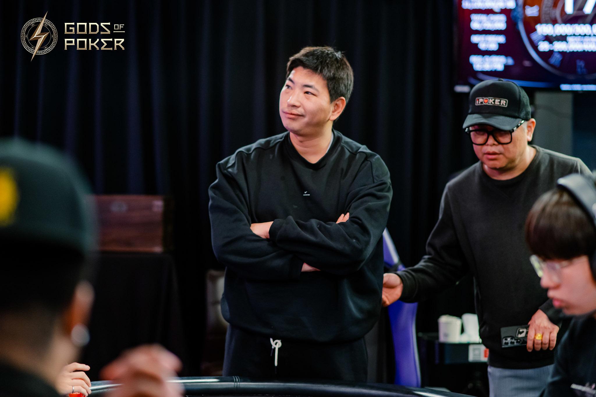 A man in black, arms crossed, intently observes a poker game.