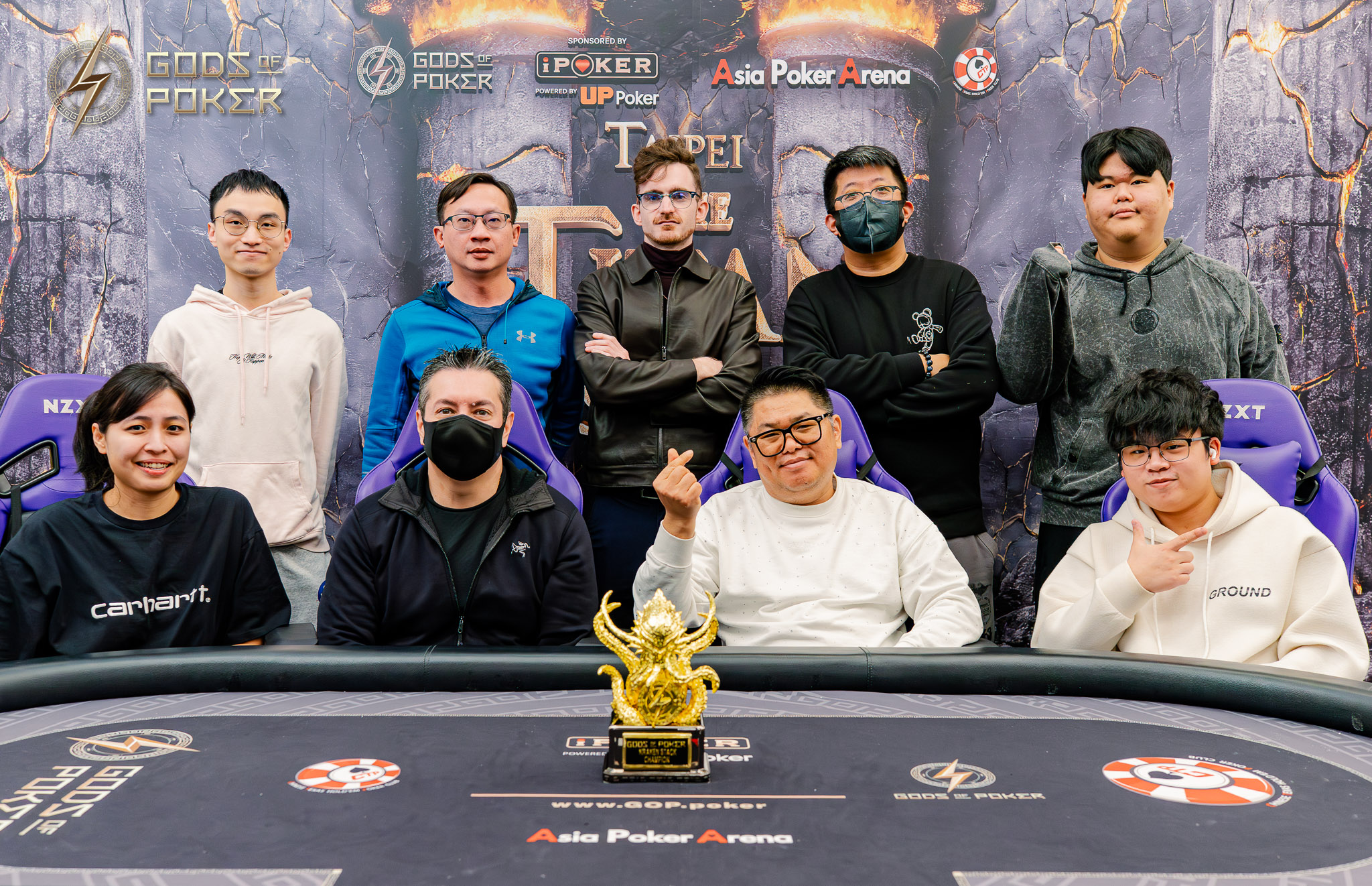 Ten poker players pose together around a poker table with a large golden trophy.