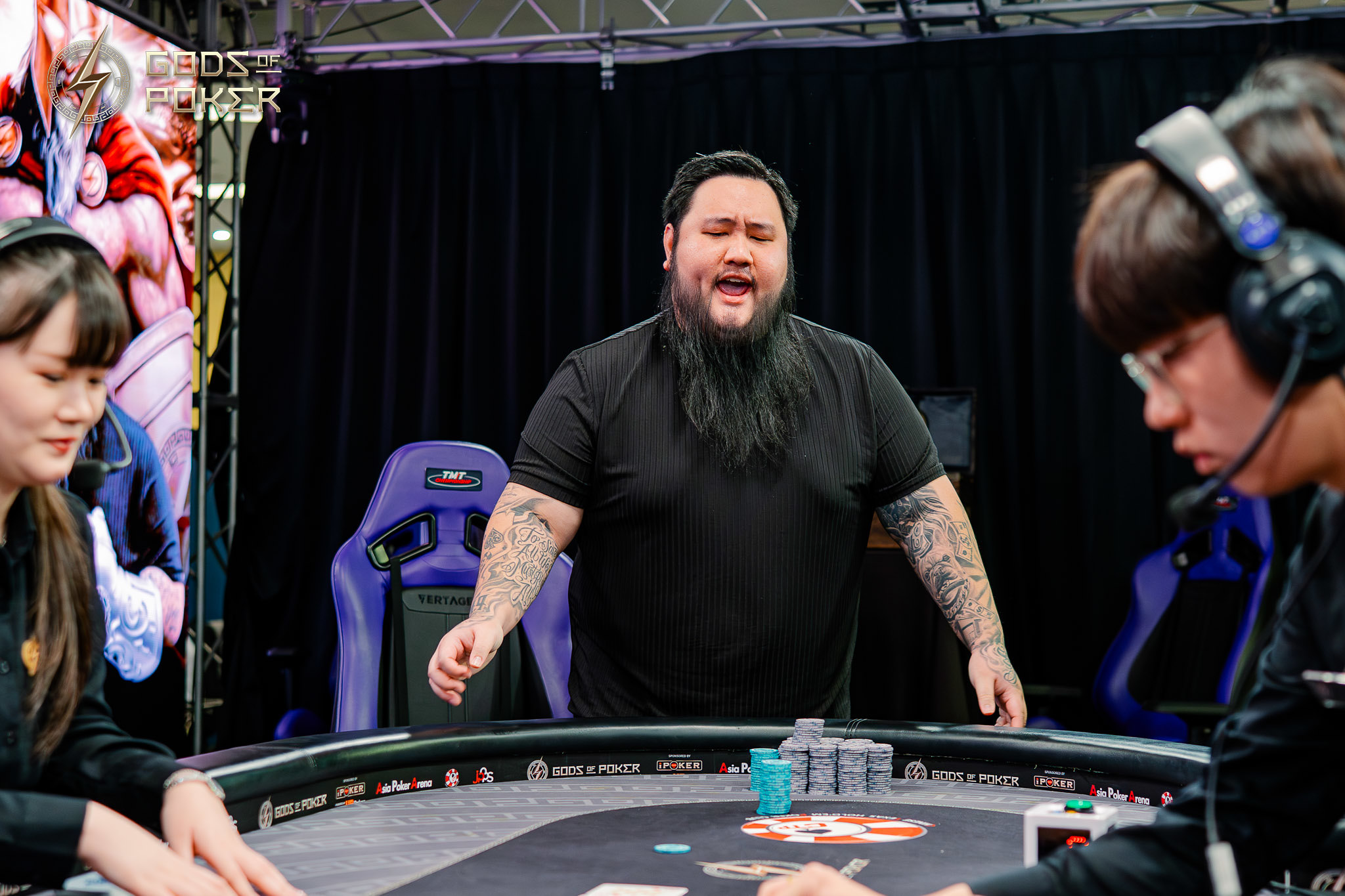 A bearded man with tattooed arms reacts animatedly at a poker table during a game.