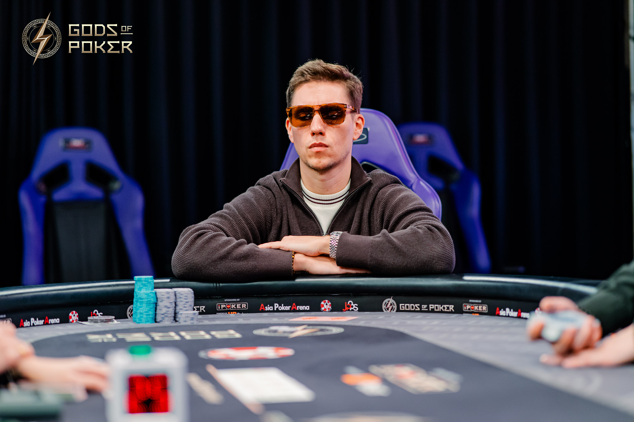 A man in sunglasses and a hoodie sits intently at a poker table with chips.