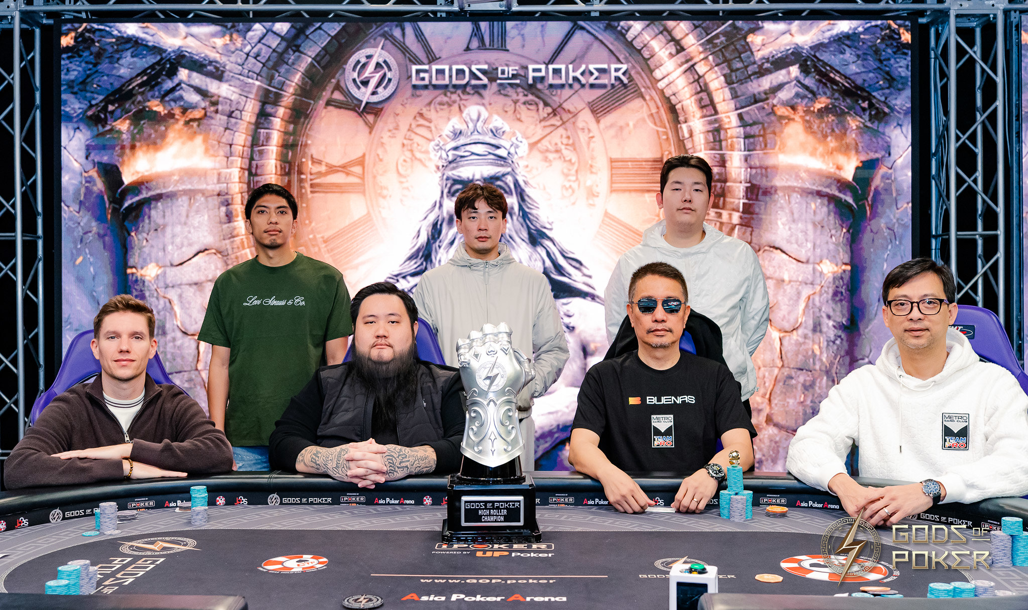 Seven men pose around a poker table with a large silver gauntlet trophy.