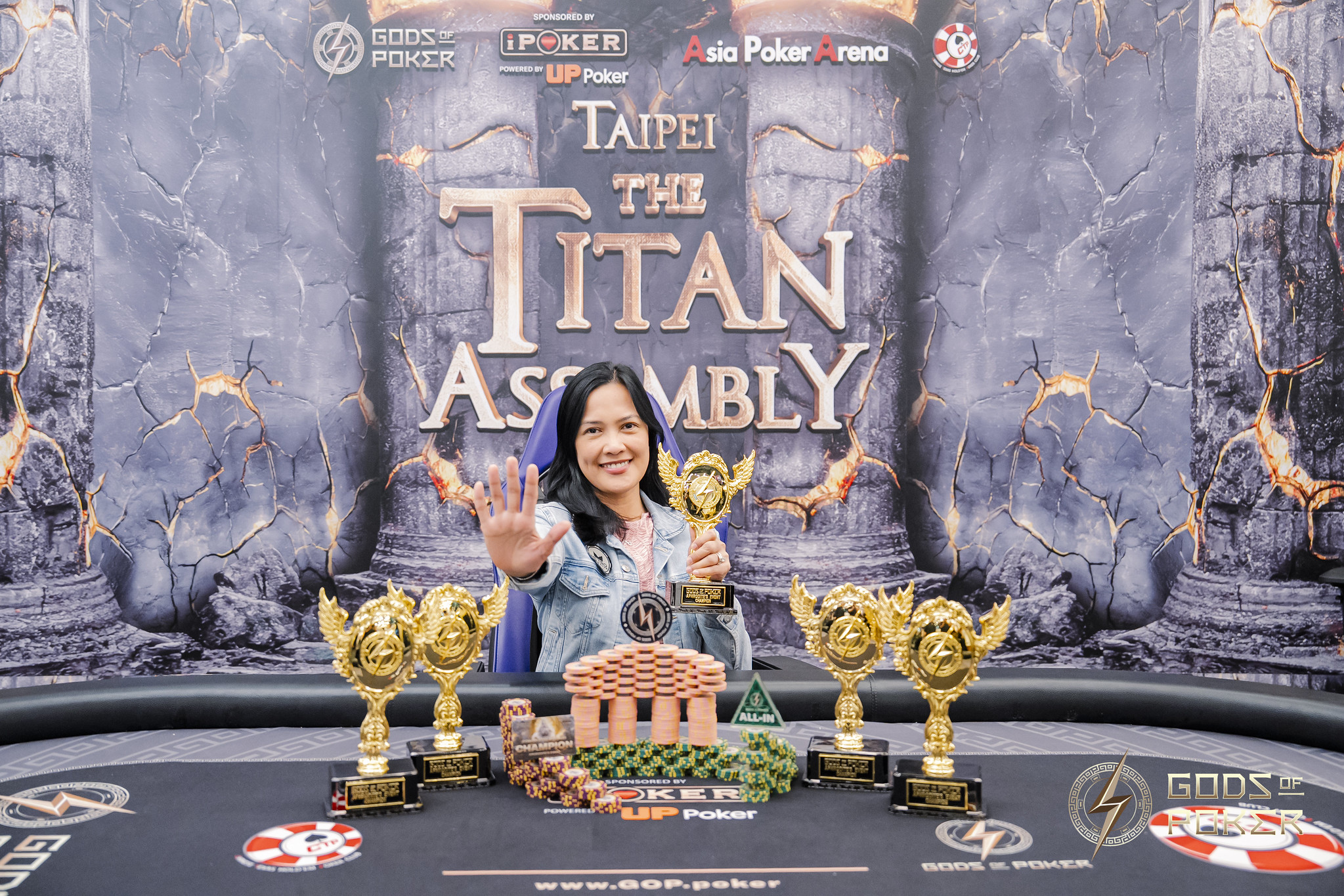 Smiling woman displays hand, surrounded by poker chips, trophies, and a miniature building.