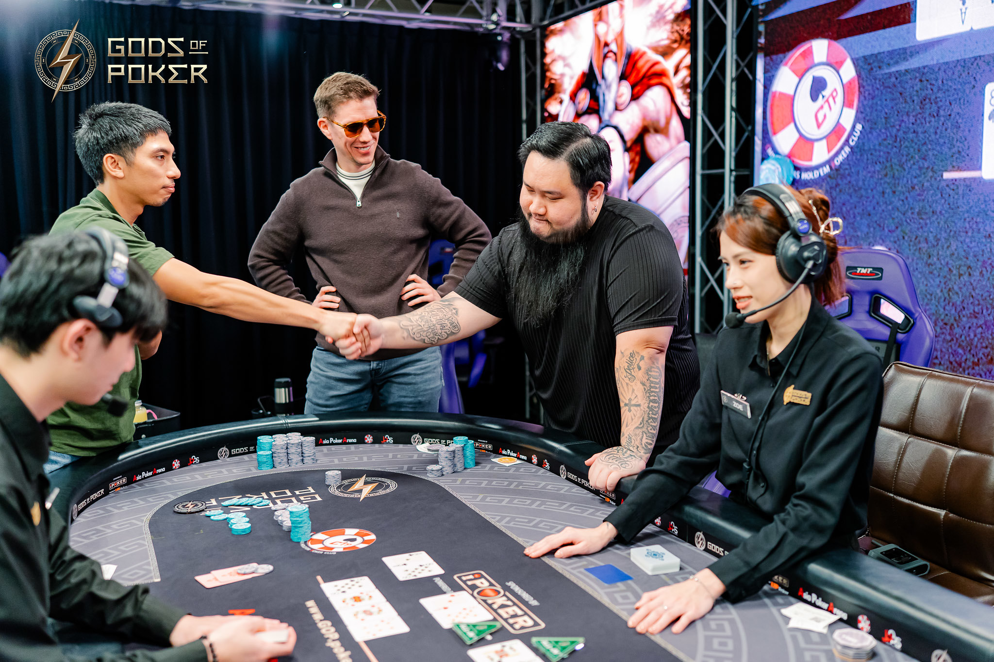 Two men shake hands over a poker table, celebrating victory at a 'Gods of Poker' event.