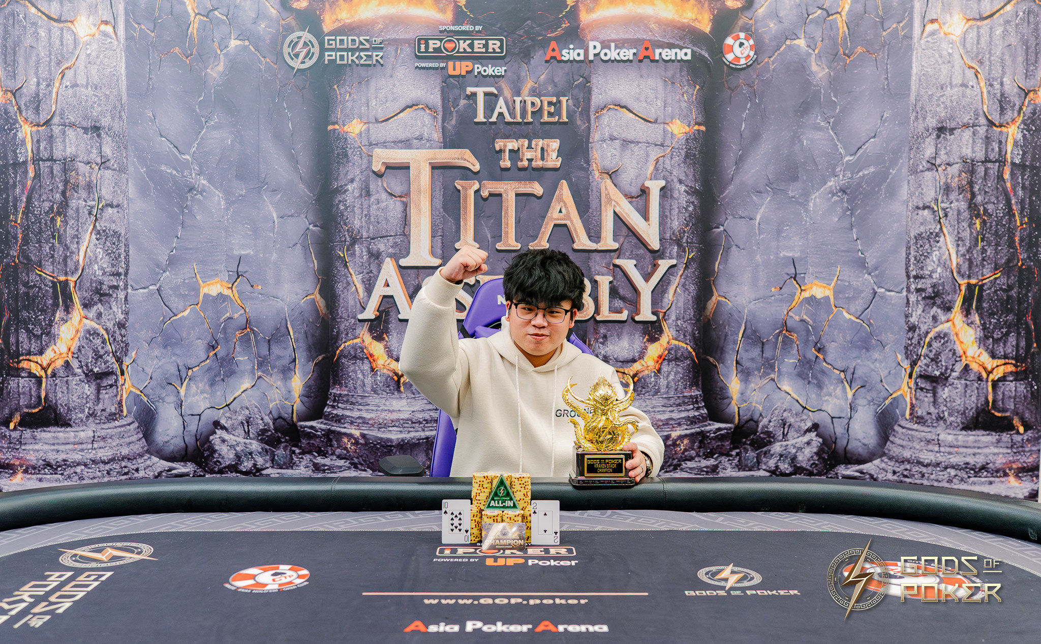 Man in hoodie celebrates poker win with golden trophy at 'The Titan Assembly' event.