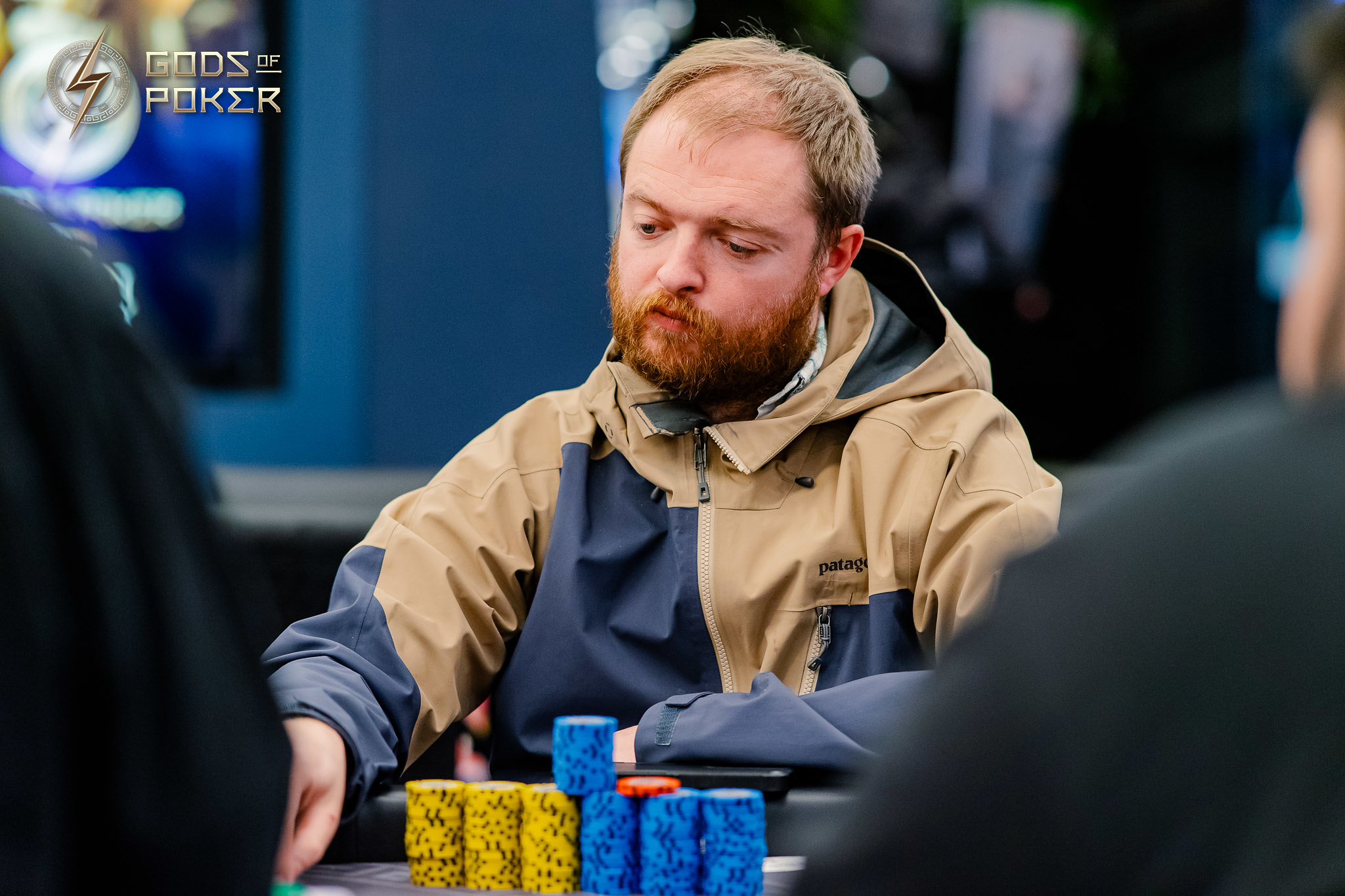A bearded man in a two-tone jacket focused at a poker table with chips.