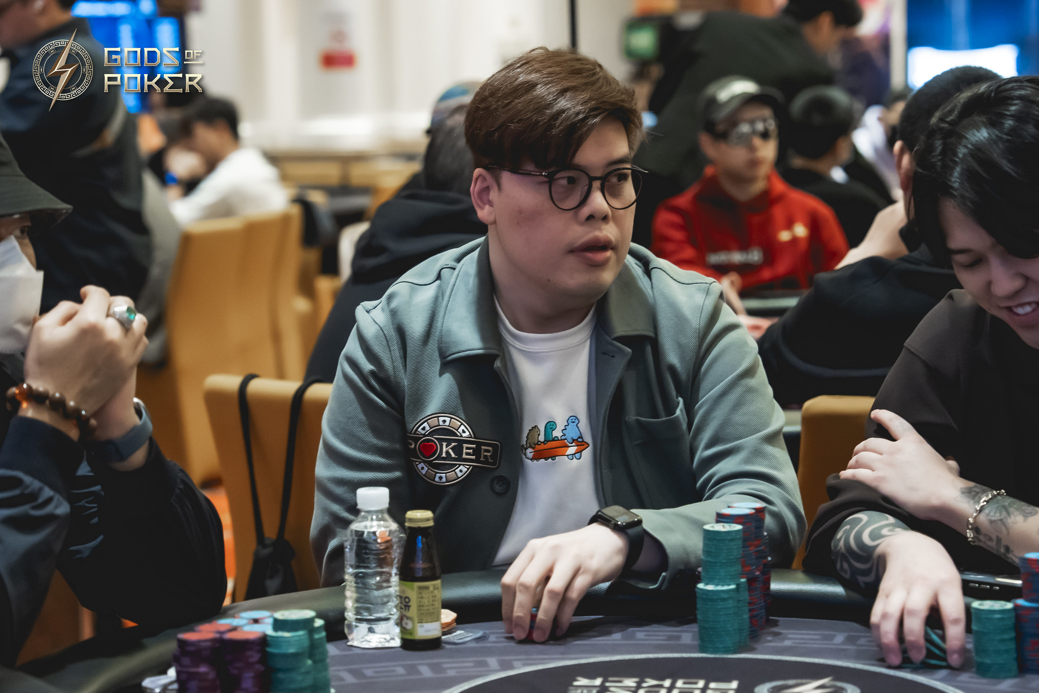 A man in glasses and a teal jacket sits intently at a poker table with chips.