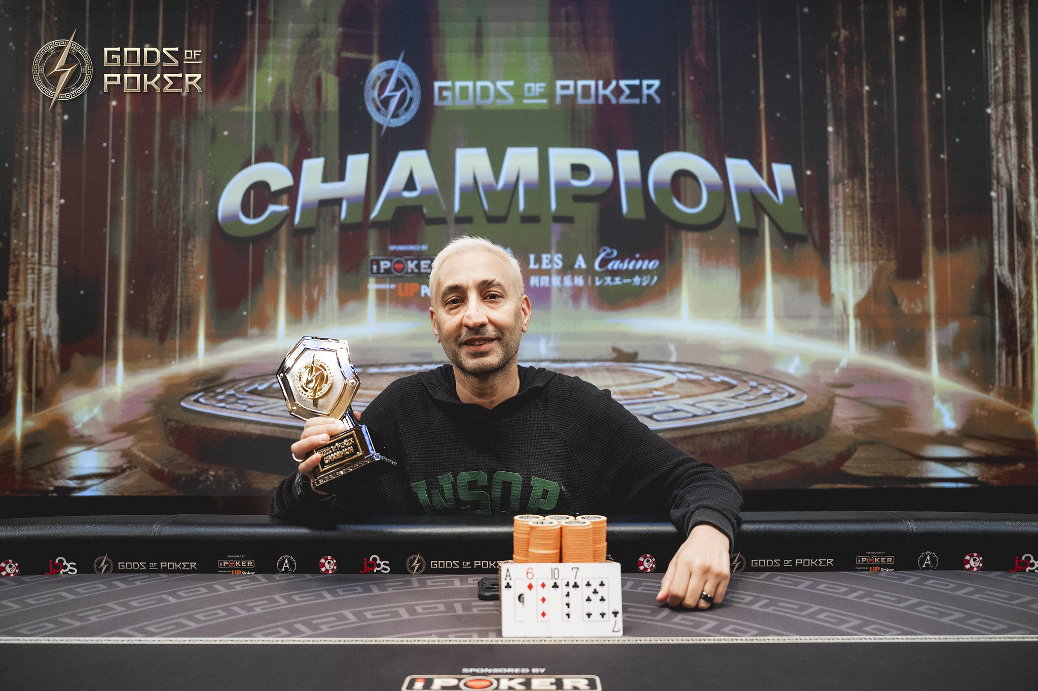 Smiling man holds a poker trophy at a table with a 'CHAMPION' screen.