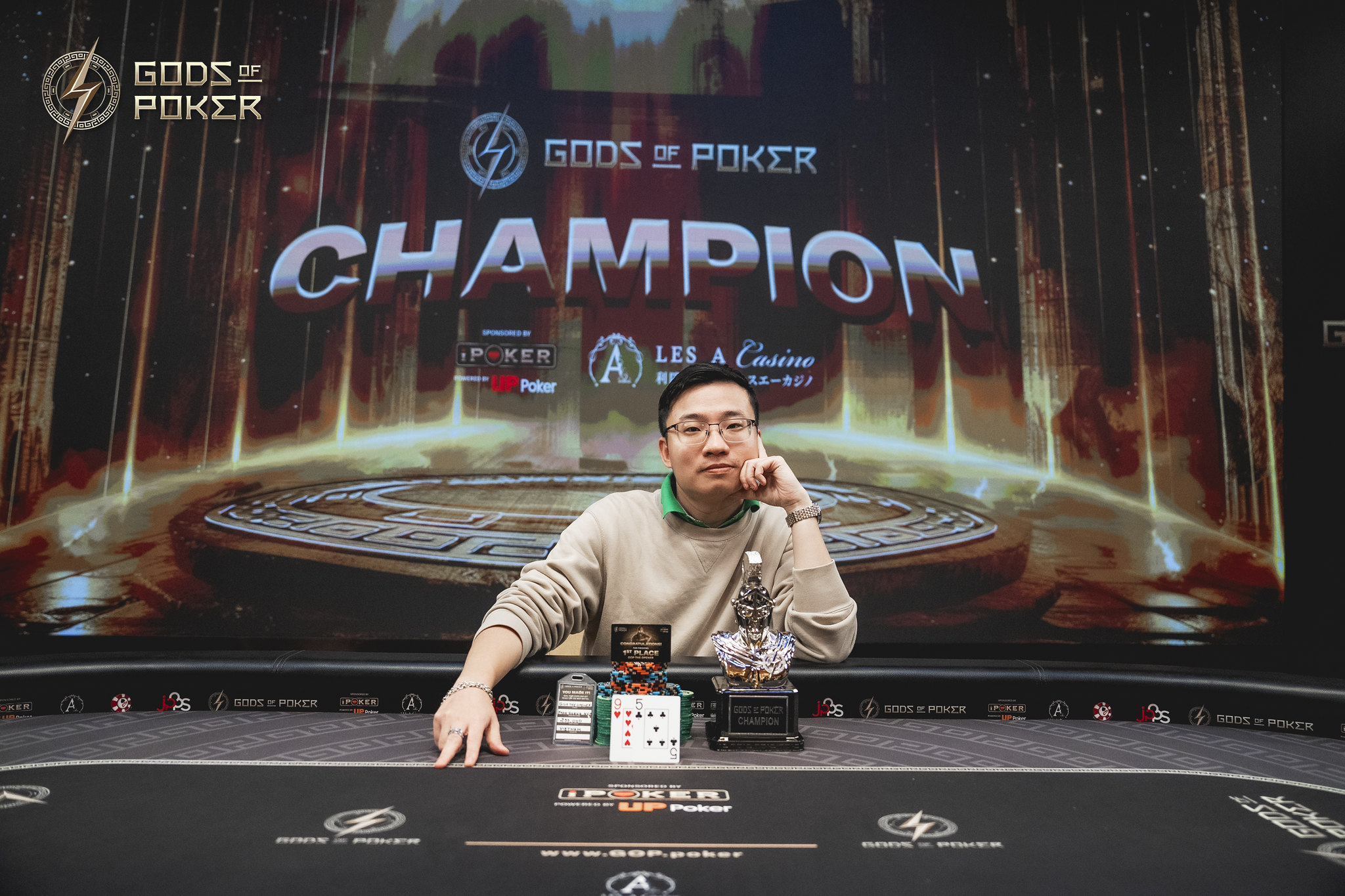 A man wearing glasses smiles behind a poker table with a championship trophy.