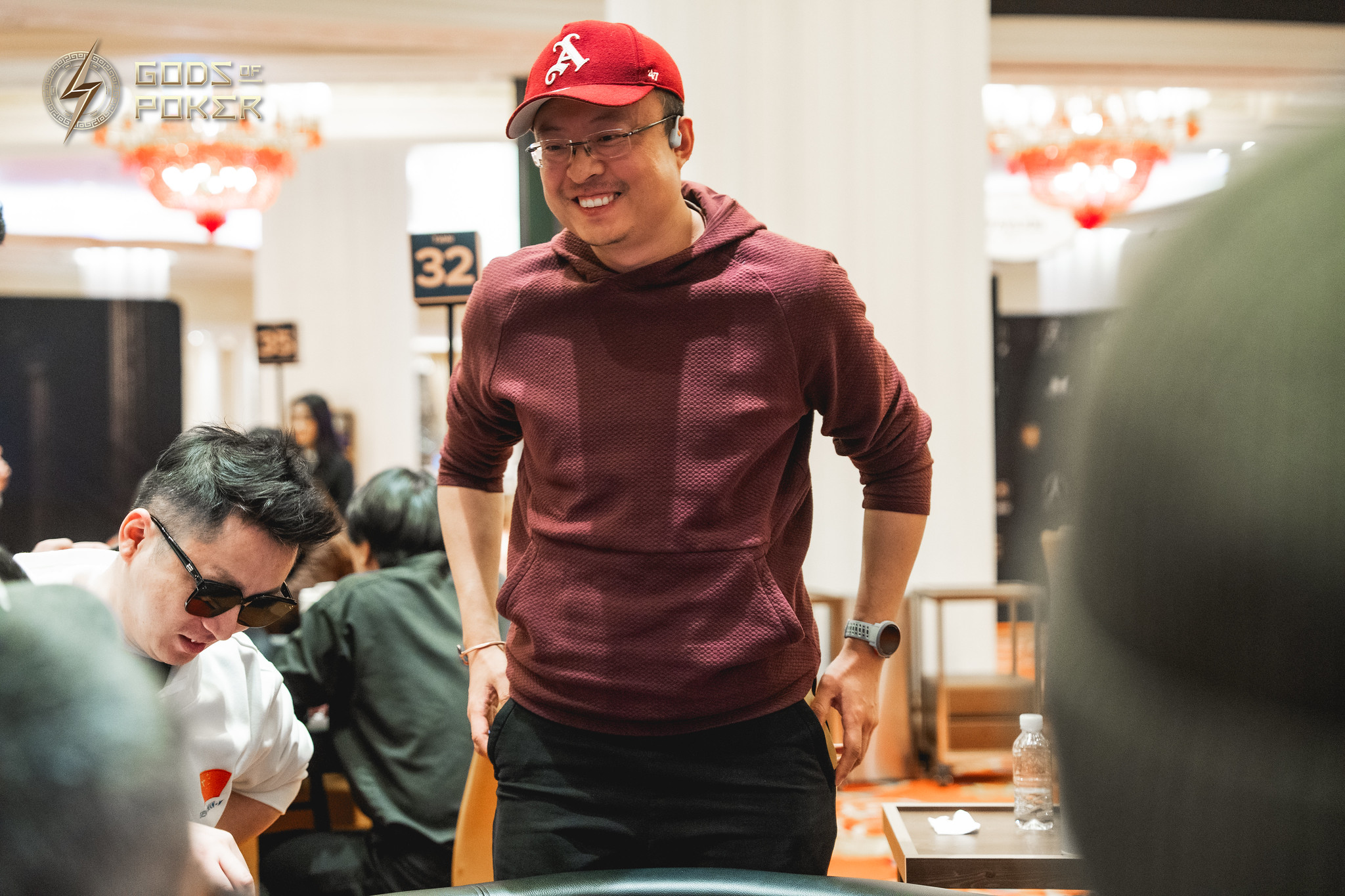 Smiling man in red cap and maroon hoodie stands by a poker table.