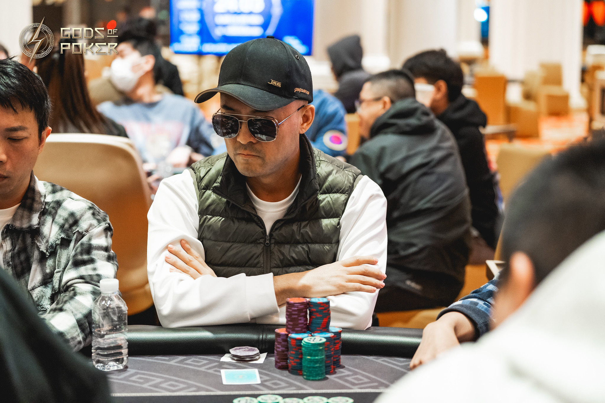 Man in cap and sunglasses sits at a poker table with chips, arms crossed.