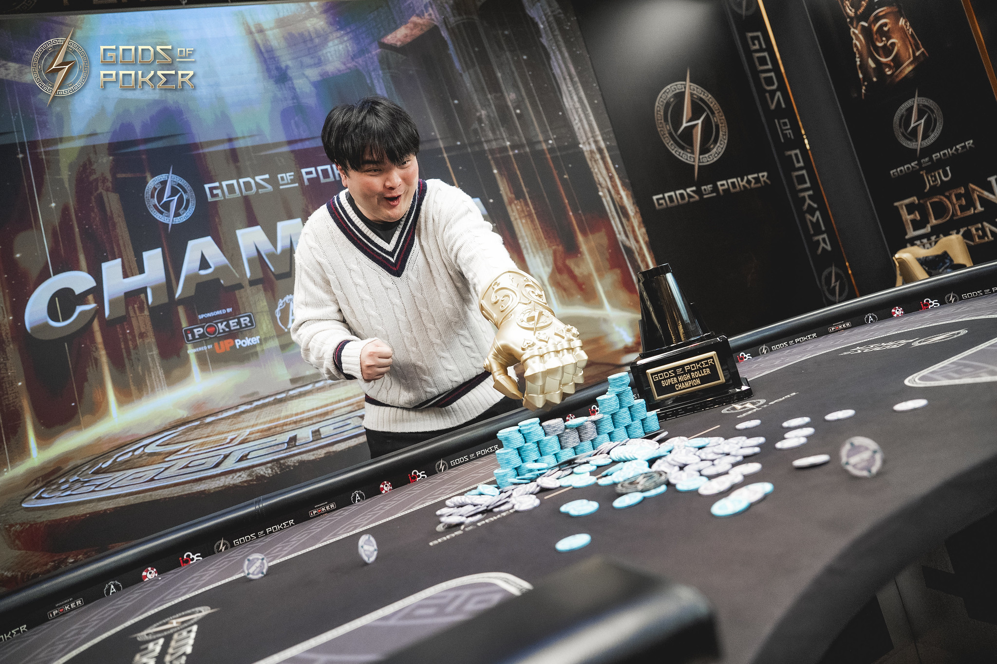 Man in white sweater celebrates poker win, sweeping blue chips towards him with a trophy nearby.