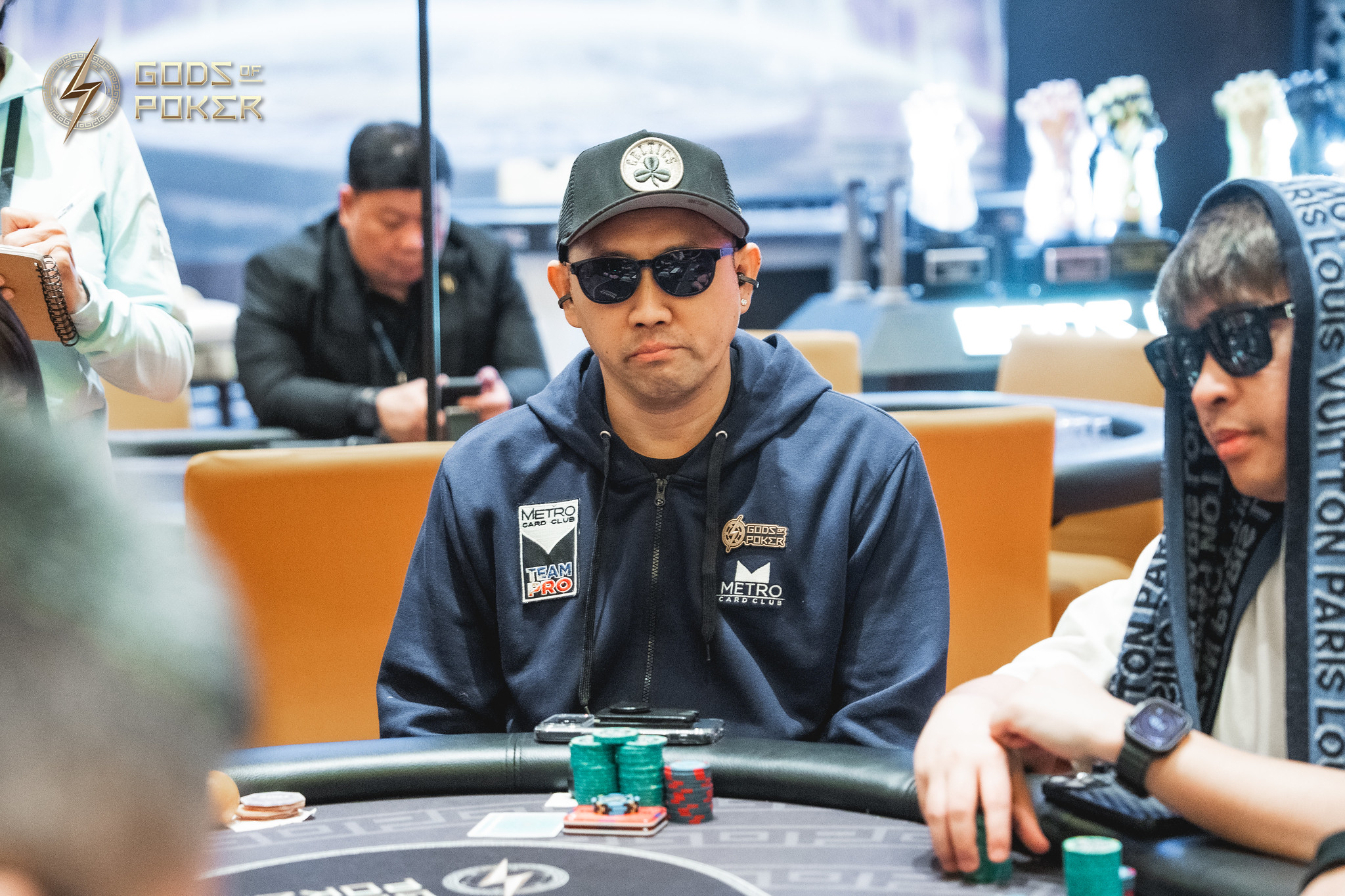 A man in a cap and sunglasses sits intensely at a poker table with chips.