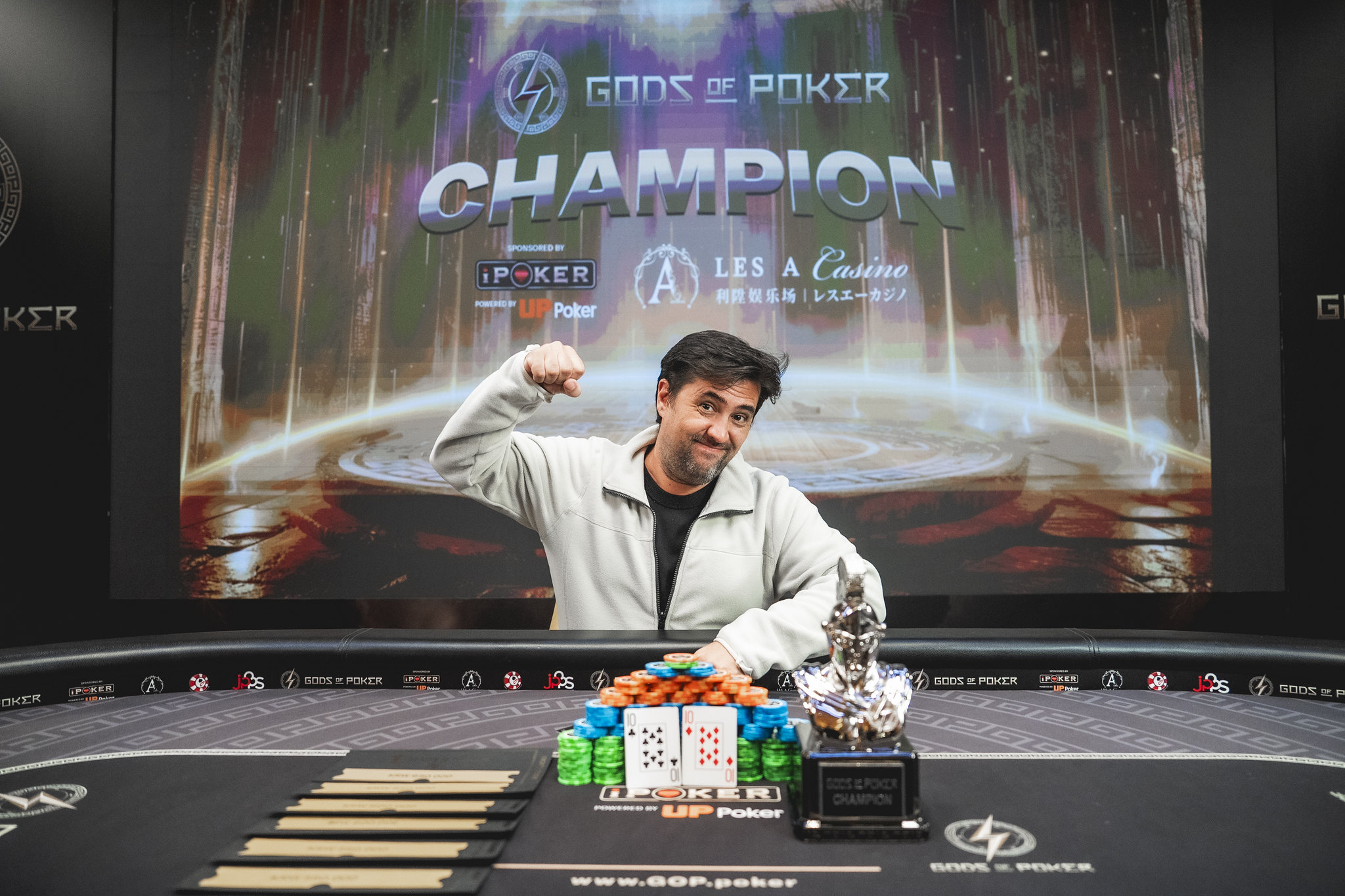 Smiling man celebrates poker championship victory with trophy and chips at the table.
