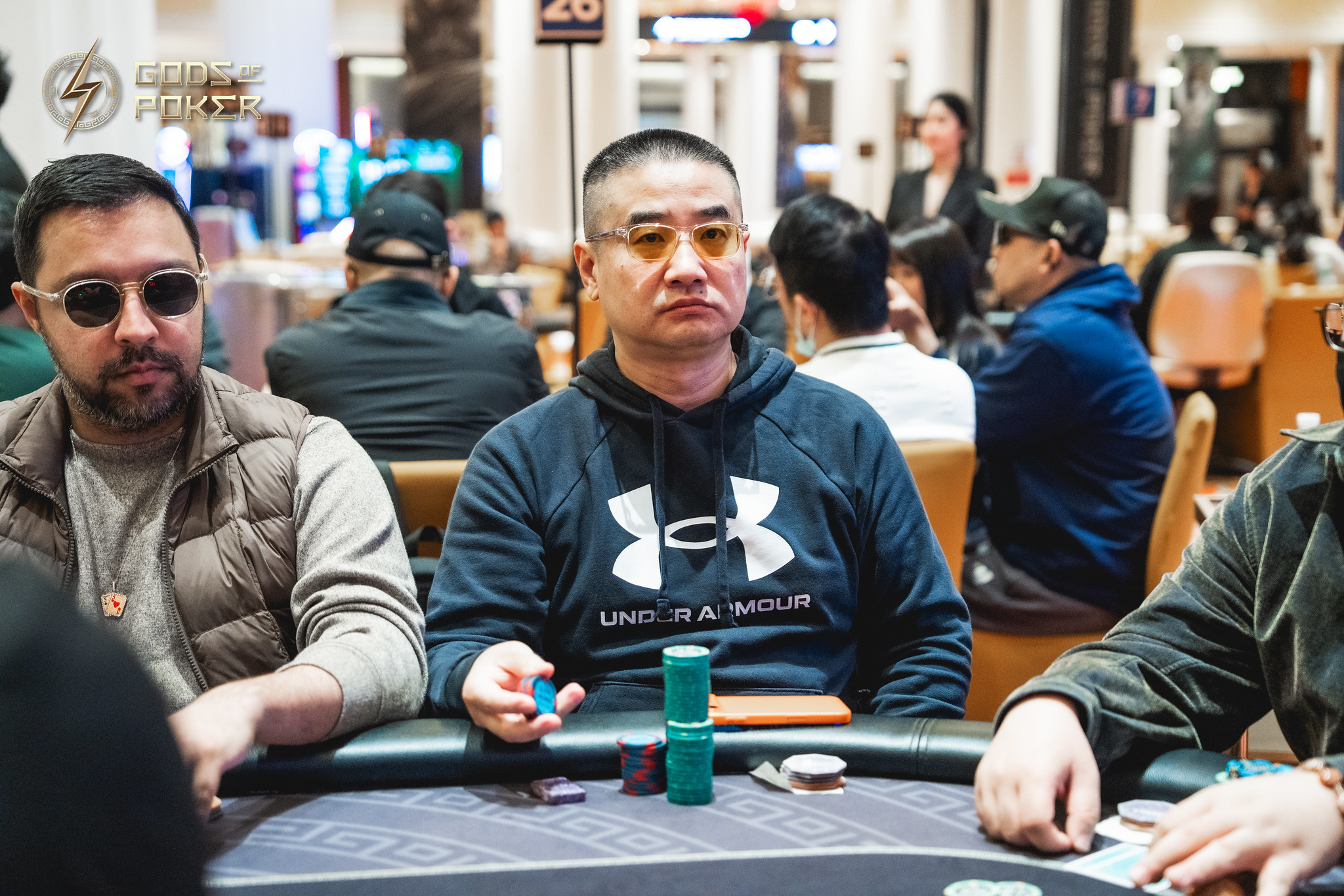 Two men seated at a poker table in a casino, one holds blue chips.