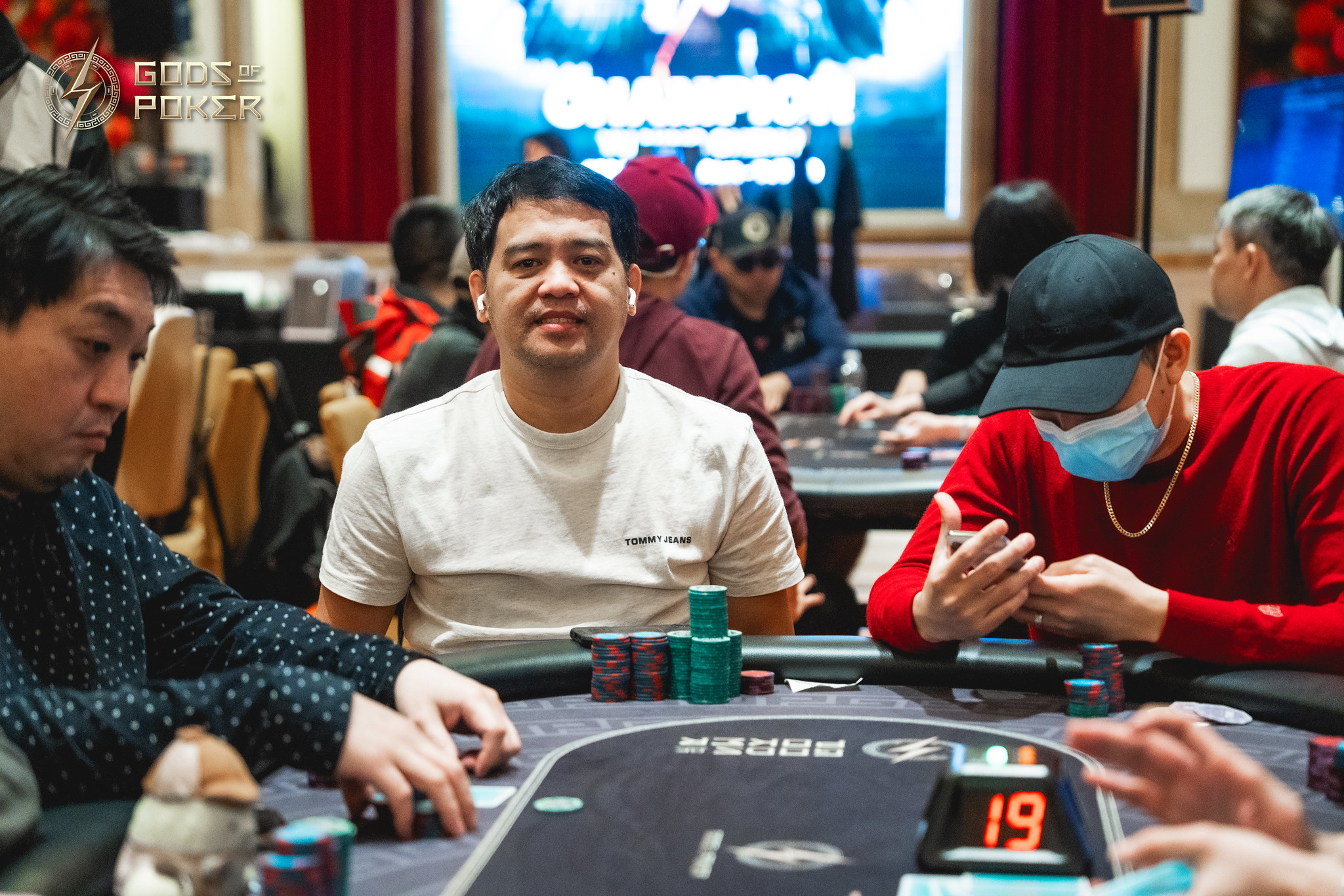 A man in a light t-shirt sits at a poker table with other players and chips.