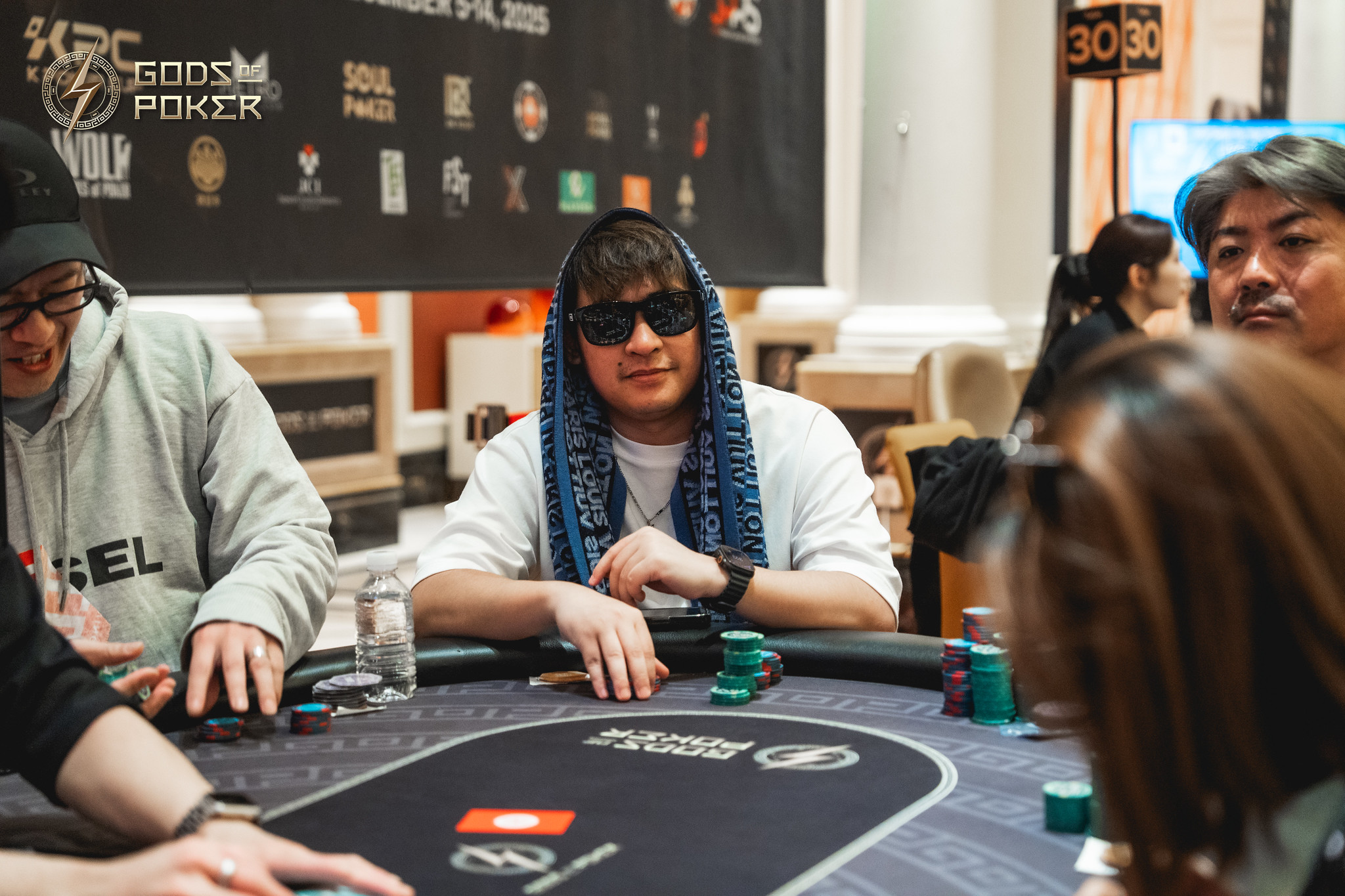 Man in sunglasses and blue scarf sitting at a poker table with chips.
