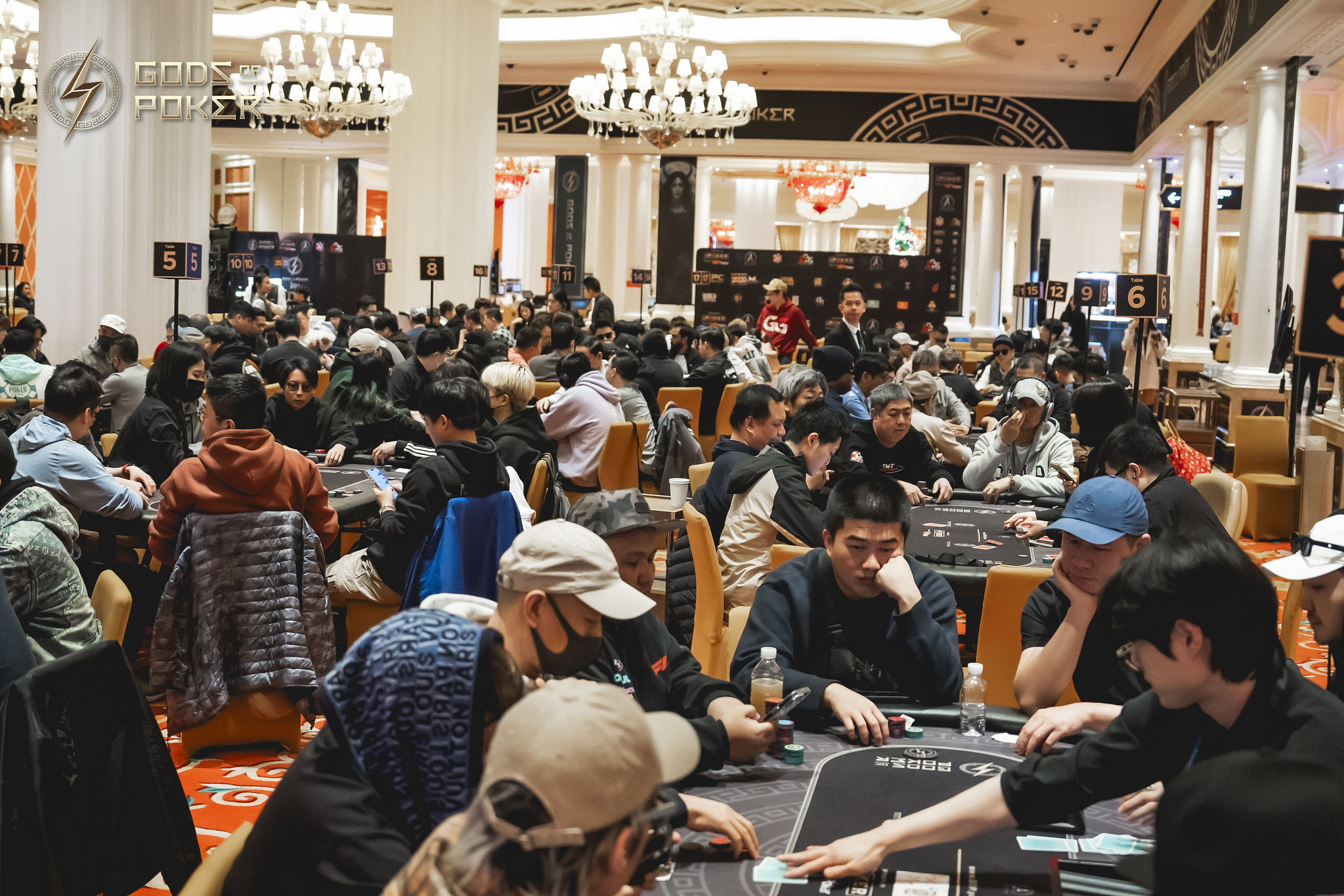 A large, ornate poker room is filled with many players actively engaged at multiple tables.