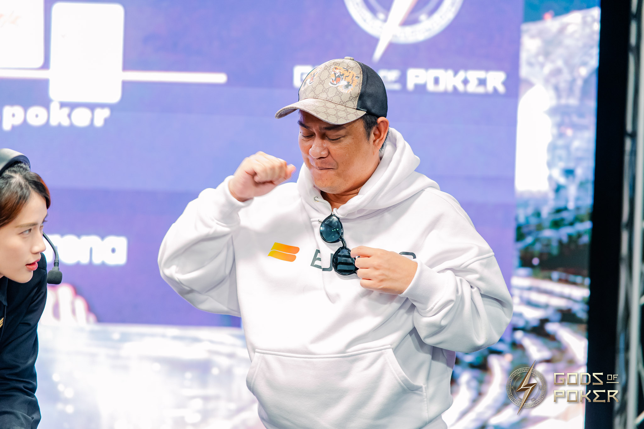 Man in a white hoodie and cap adjusting his clothing at a poker event.