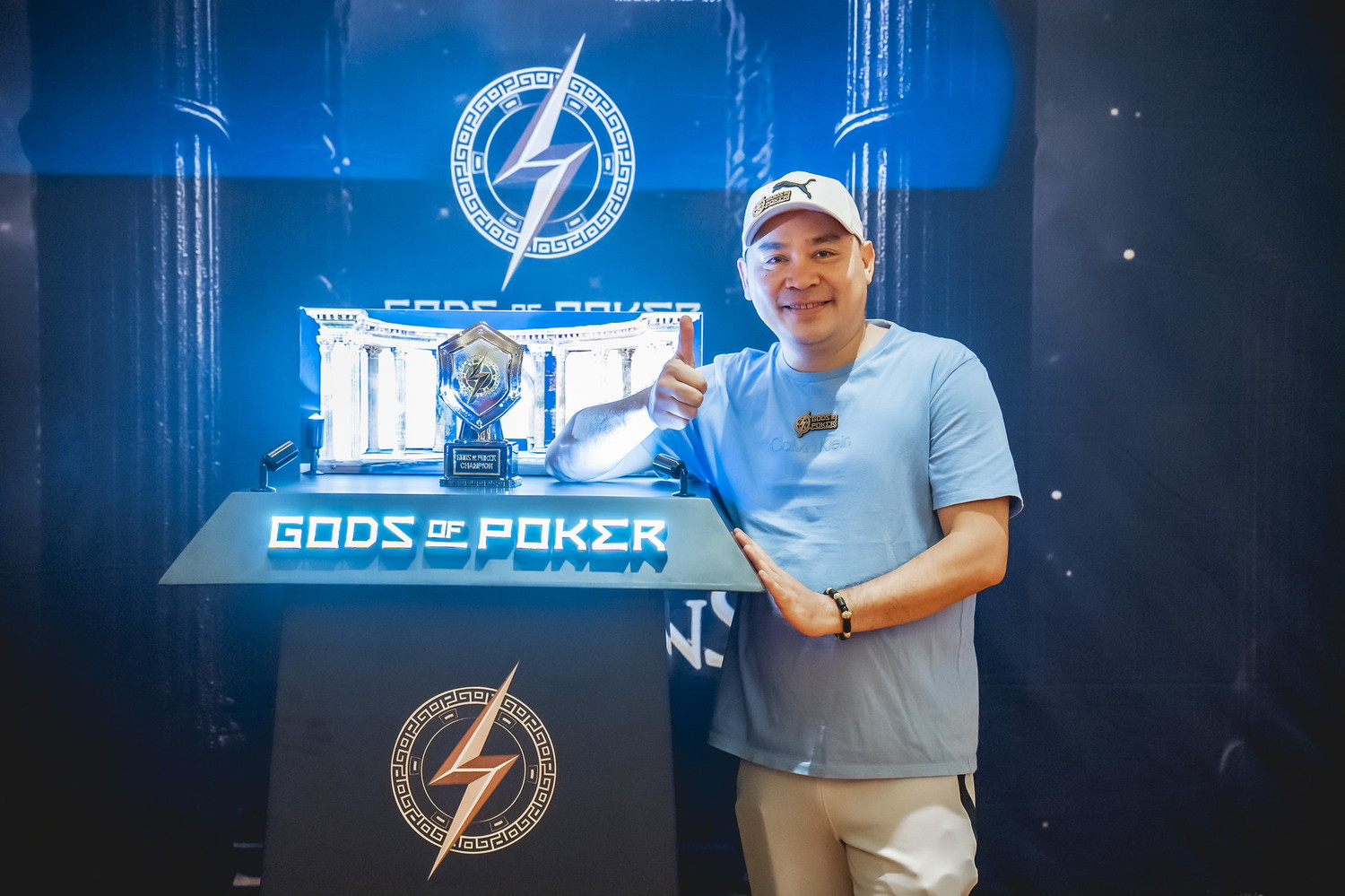Man gives thumbs up next to a glowing 'Gods of Poker' trophy display.