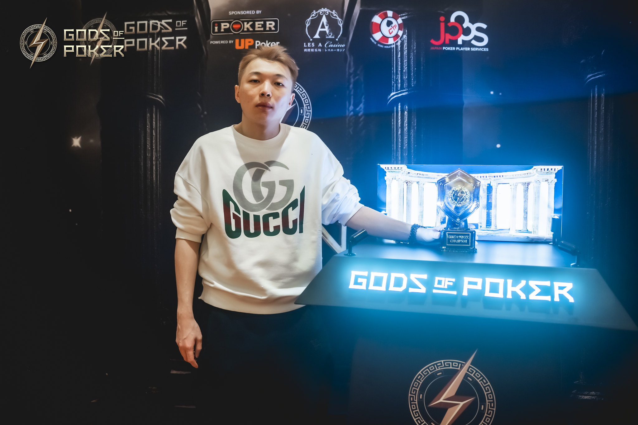 Man in Gucci sweatshirt beside a blue lit Gods of Poker display and trophy.