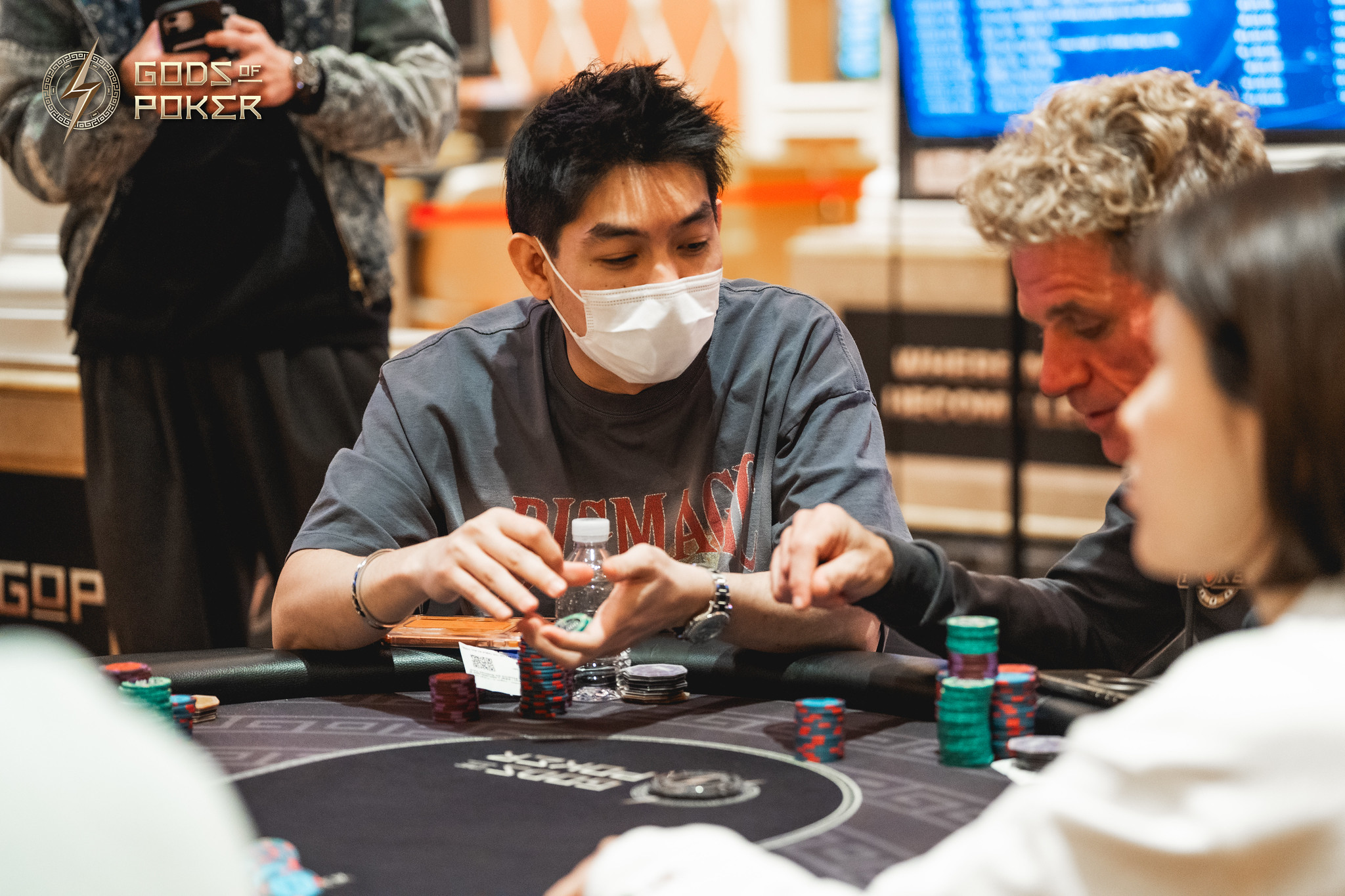 A masked man plays poker at a casino table, holding cards and chips.