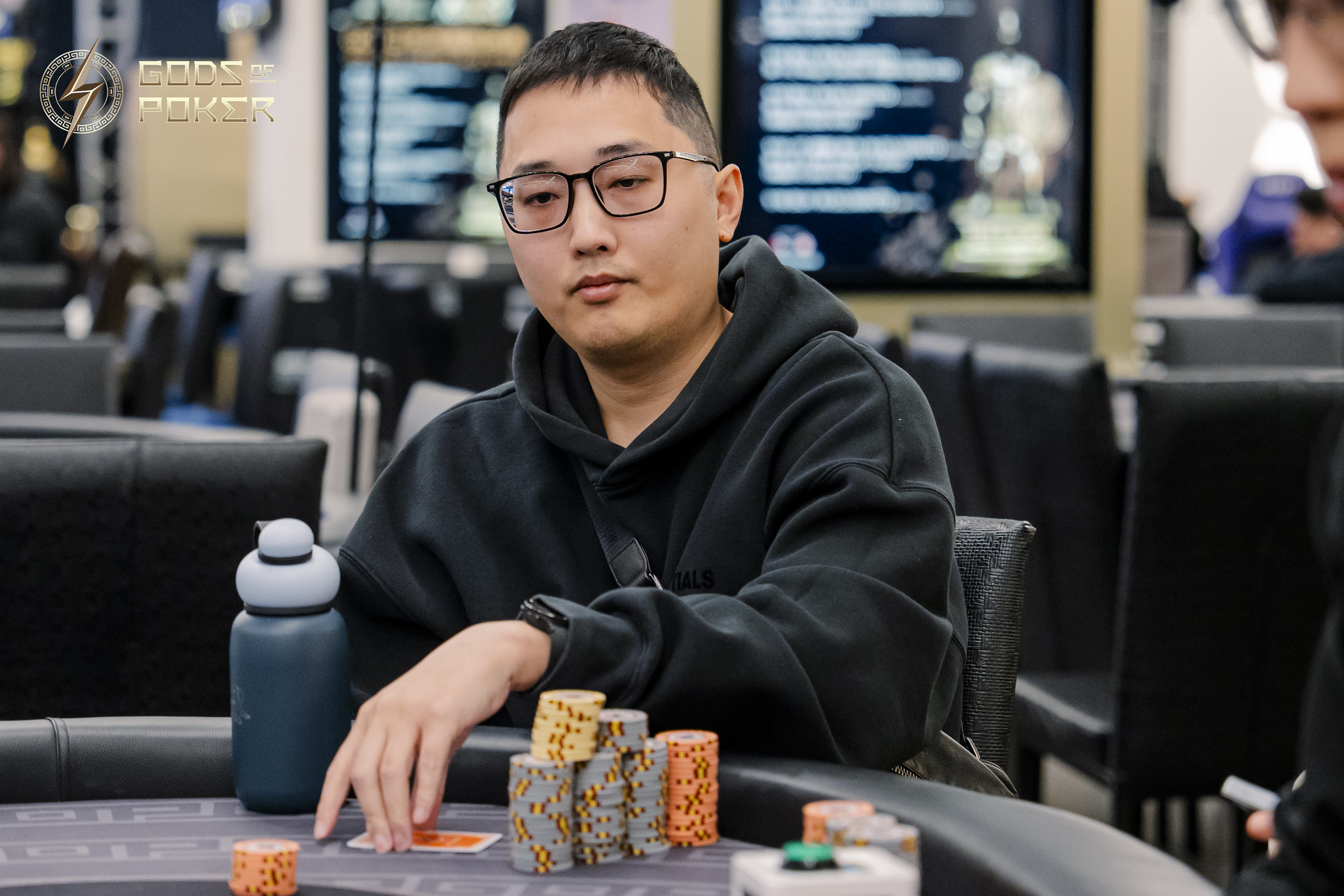 Young Asian man with glasses and black hoodie playing poker with chip stacks.
