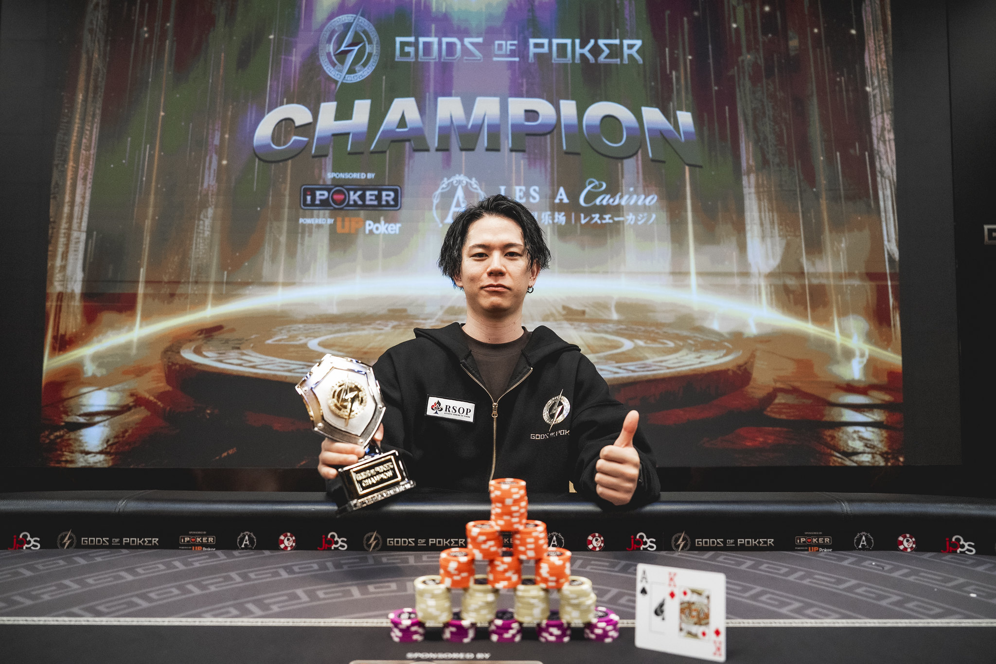 Poker champion holds a trophy and gives a thumbs-up at a poker table.