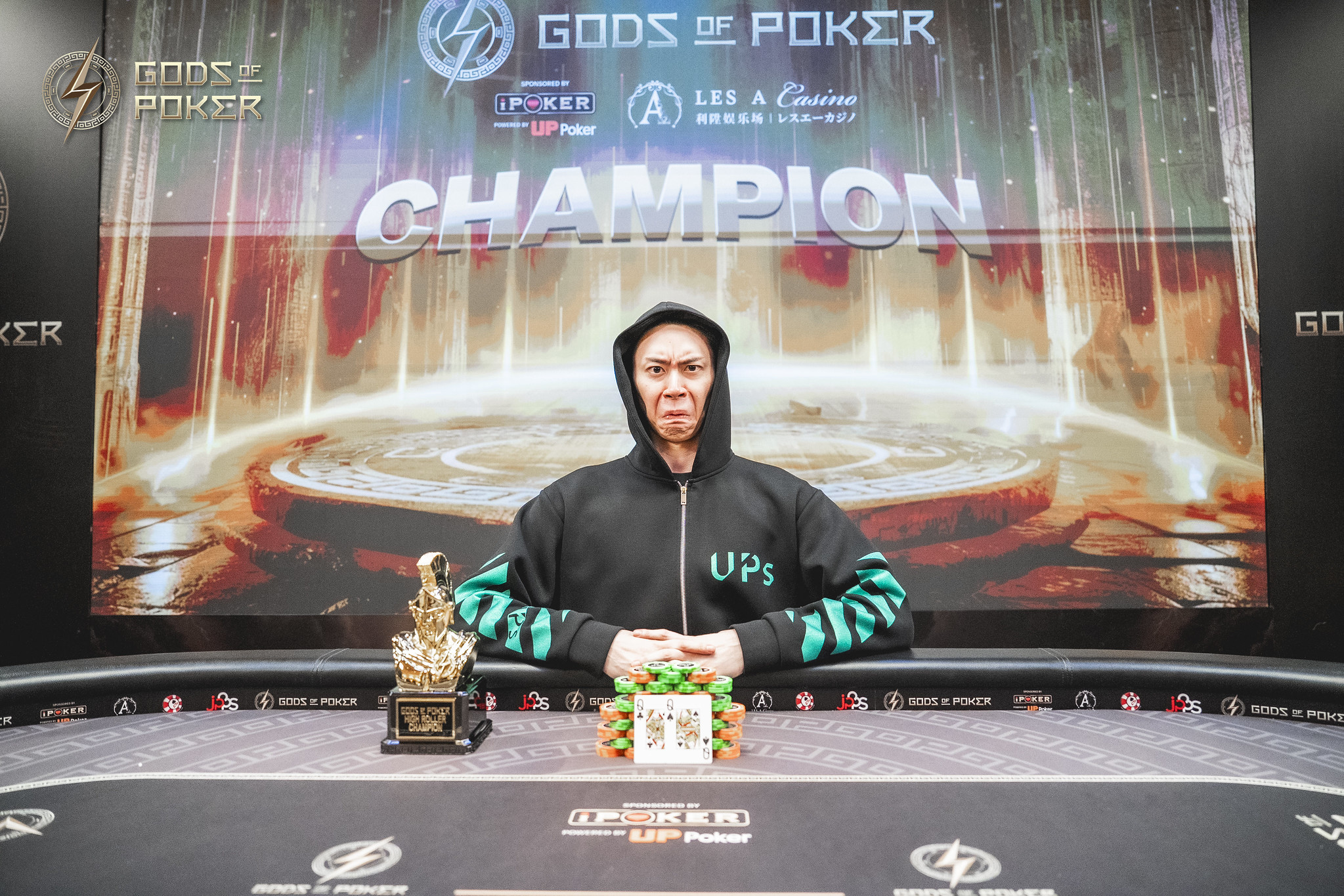 Poker champion in a black hoodie sits at a table with a trophy and chips.