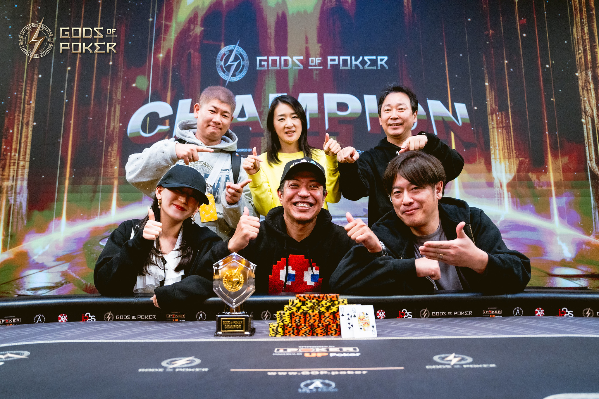 Six smiling poker champions pose with a trophy and chip stacks at a tournament.