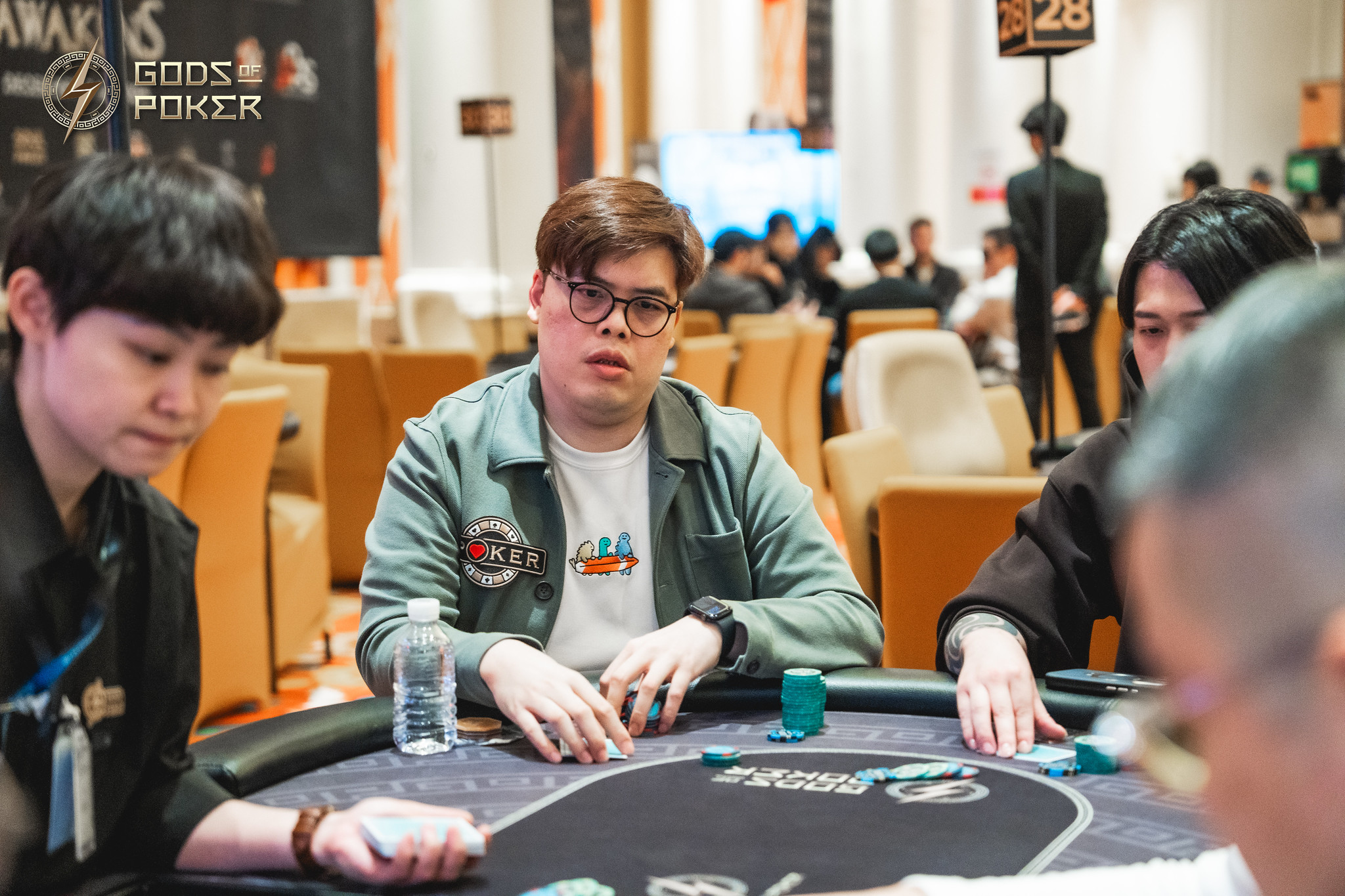 A man wearing glasses sits intently at a poker table with chips, looking forward.