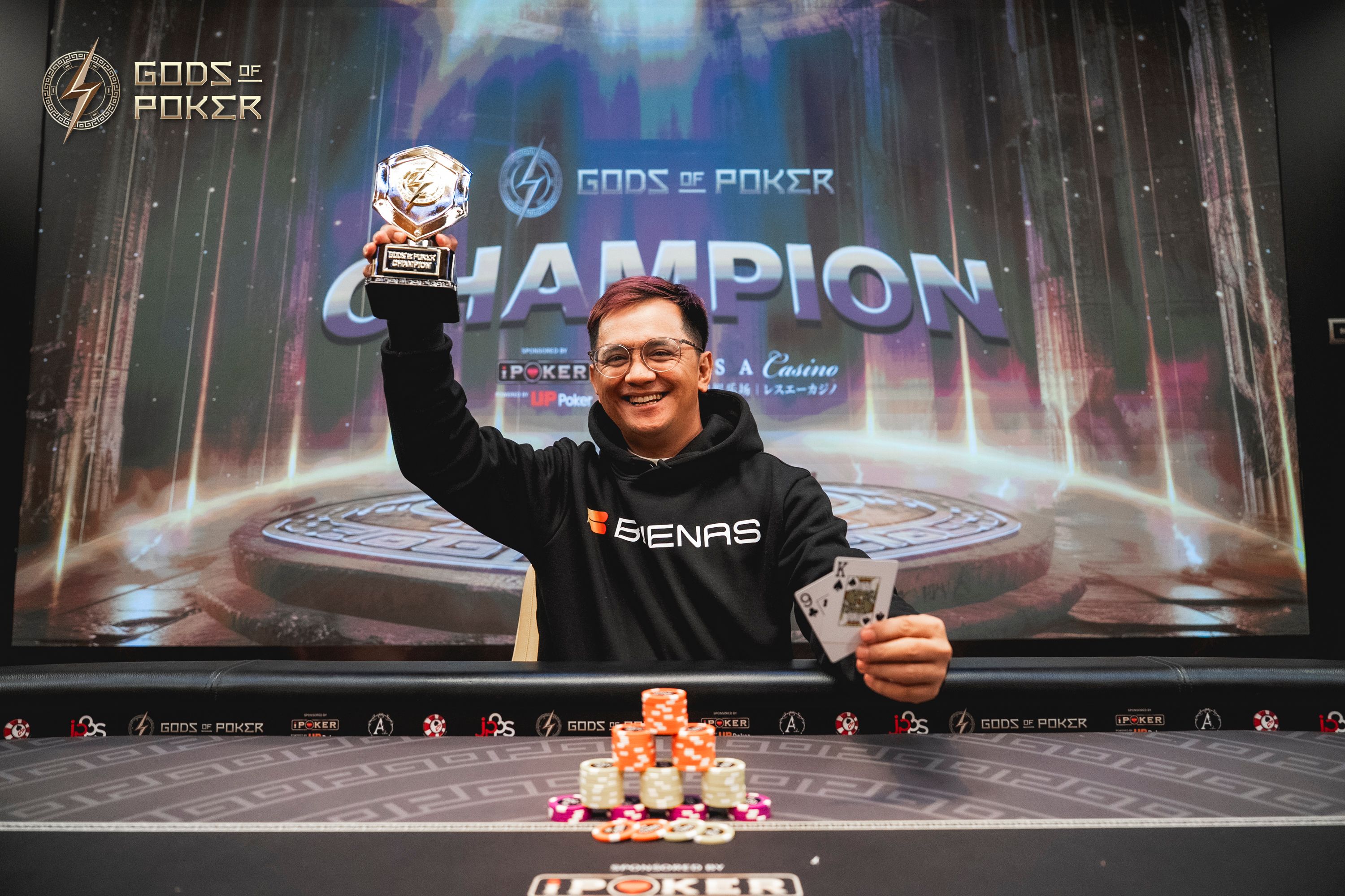 Smiling poker champion holds a trophy and winning cards in front of a large screen.