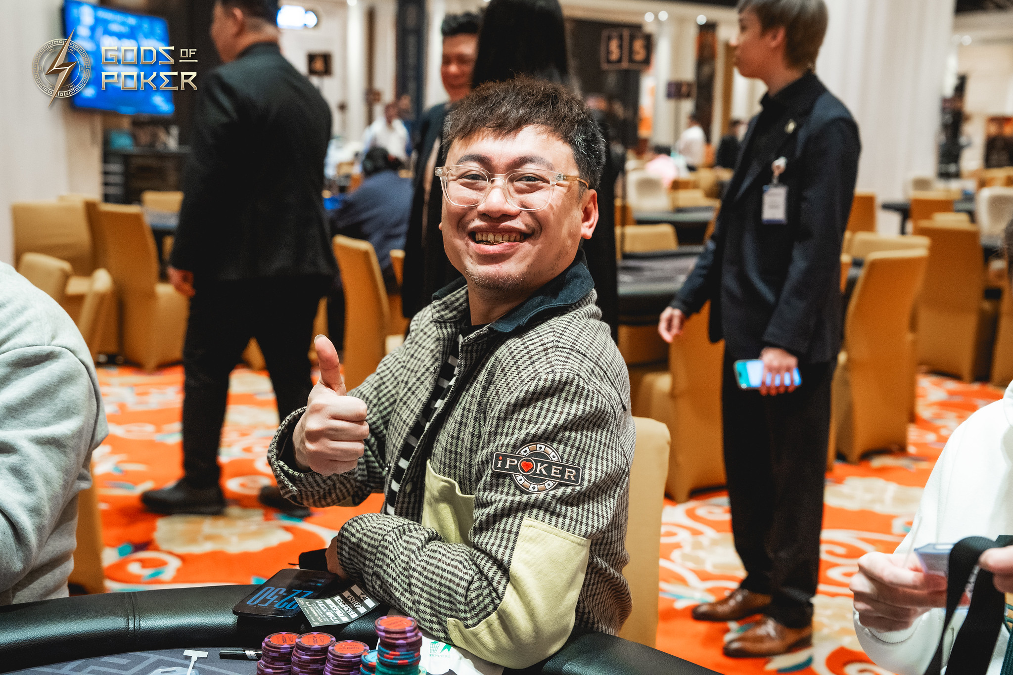 Smiling man in glasses at a poker table gives a thumbs-up.