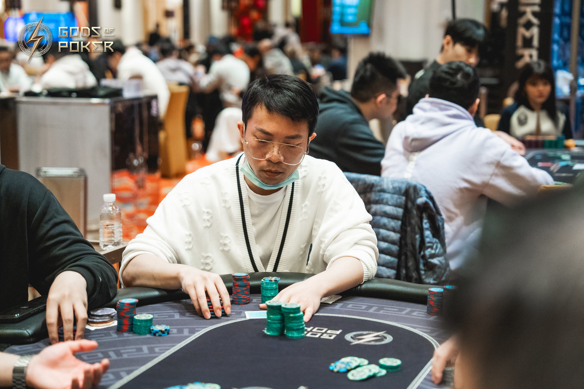 A man wearing glasses and a light sweater plays poker at a busy casino table.