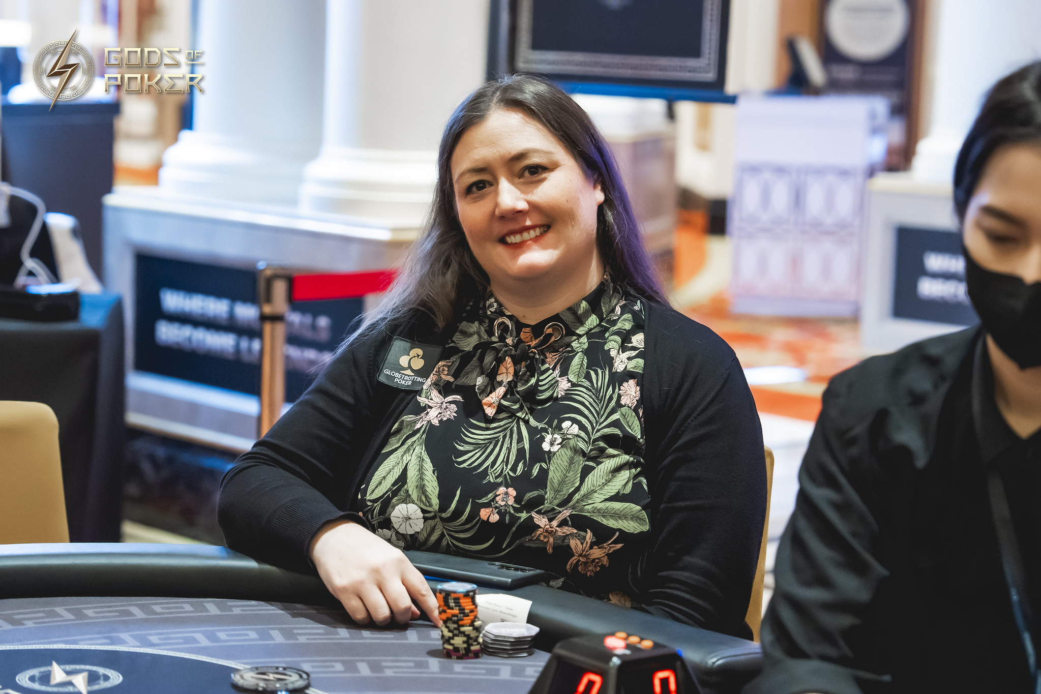 A smiling woman with dark hair sits at a poker table with chips.