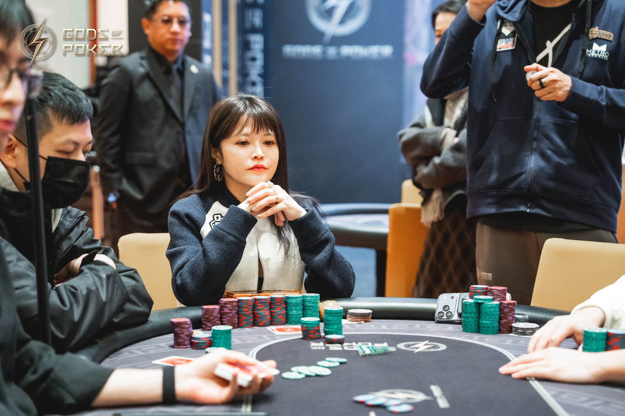 A focused woman sits at a poker table with chips, hands clasped under her chin.