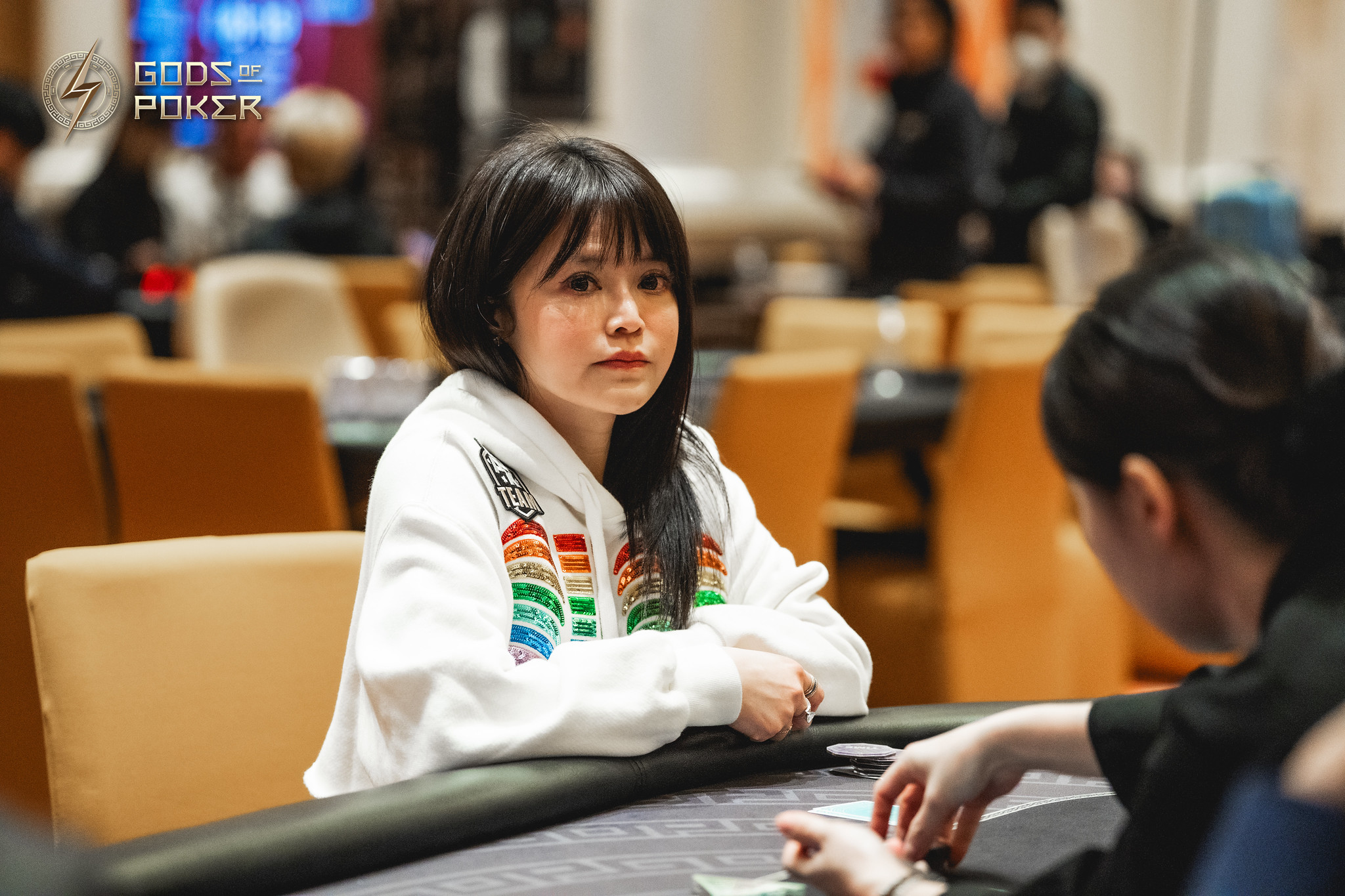A young woman in a rainbow hoodie sits intently at a poker table during a game.